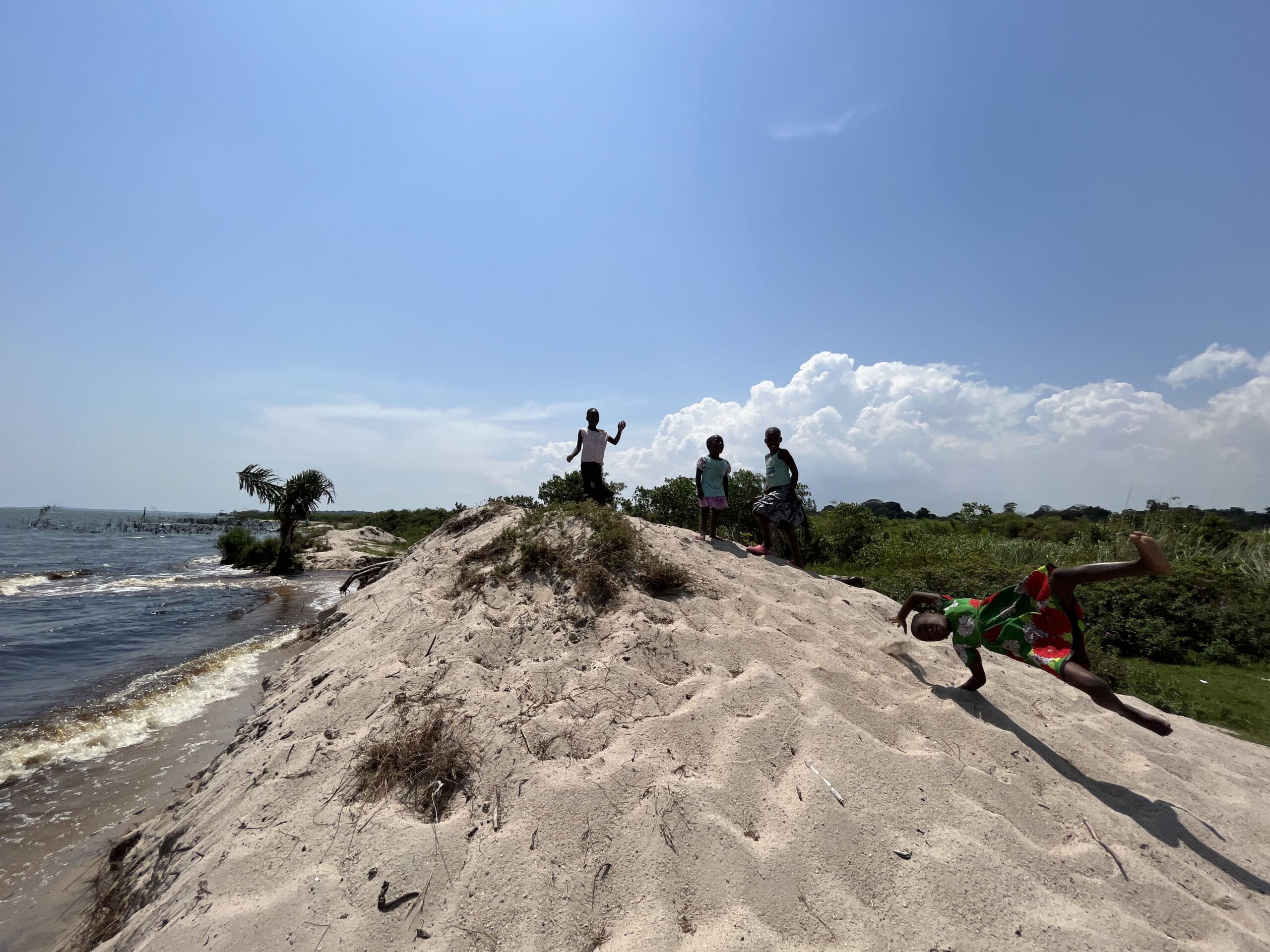 Vier kinderen spelen op een zandheuvel bij het water onder een helderblauwe hemel. Eén kind maakt een radslag op de voorgrond terwijl de anderen, als jonge reizigers, bovenop de heuvel staan, omringd door groen.