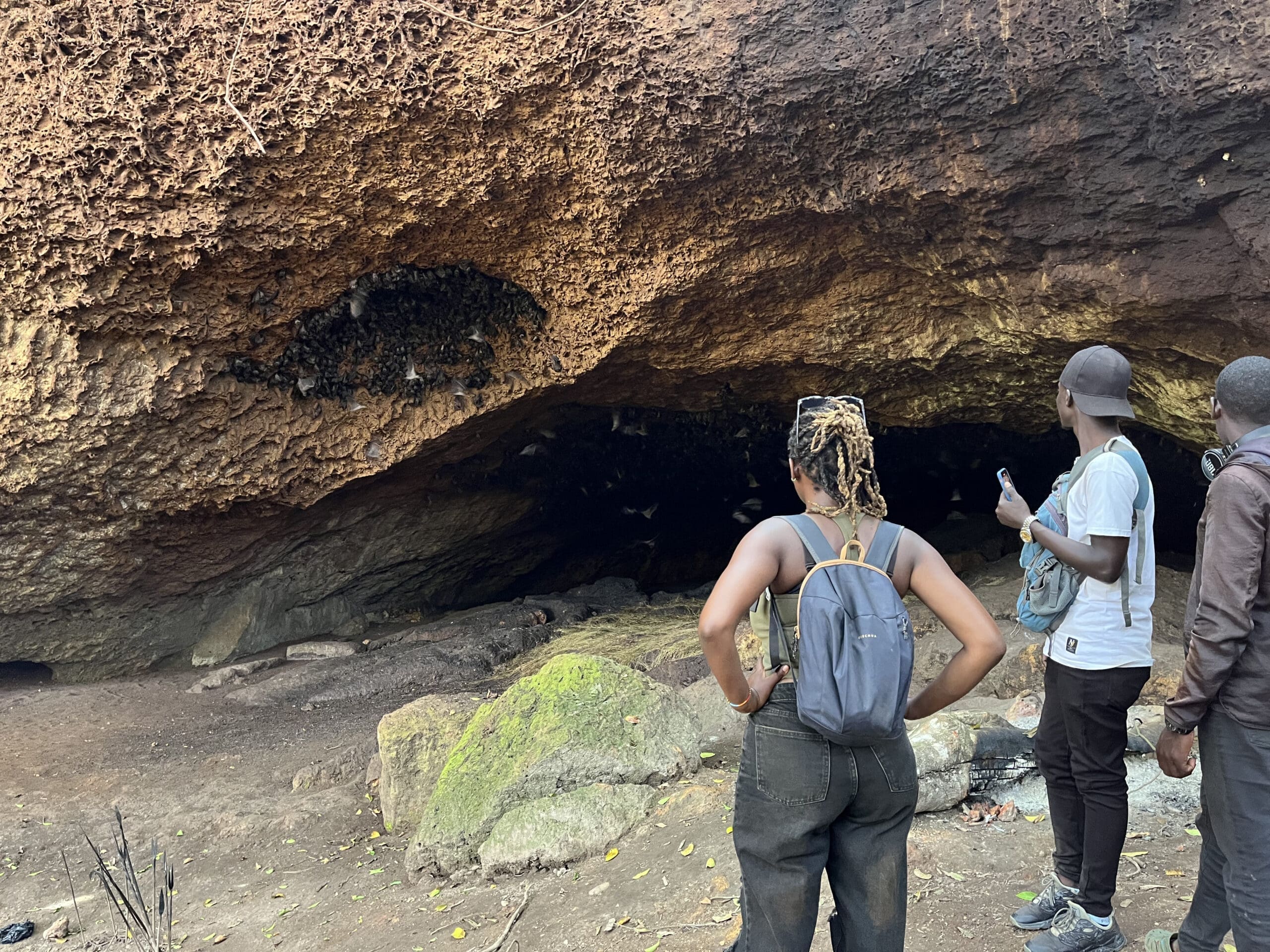 Drie mensen staan bij de ingang van een rotsachtige grot in Oeganda en staren naar binnen waar een kolonie vleermuizen zich vastklampt aan het plafond. Mosachtige rotsen en droge grond vullen de voorgrond en geven de geest van reizen en avontuur in Afrika weer.