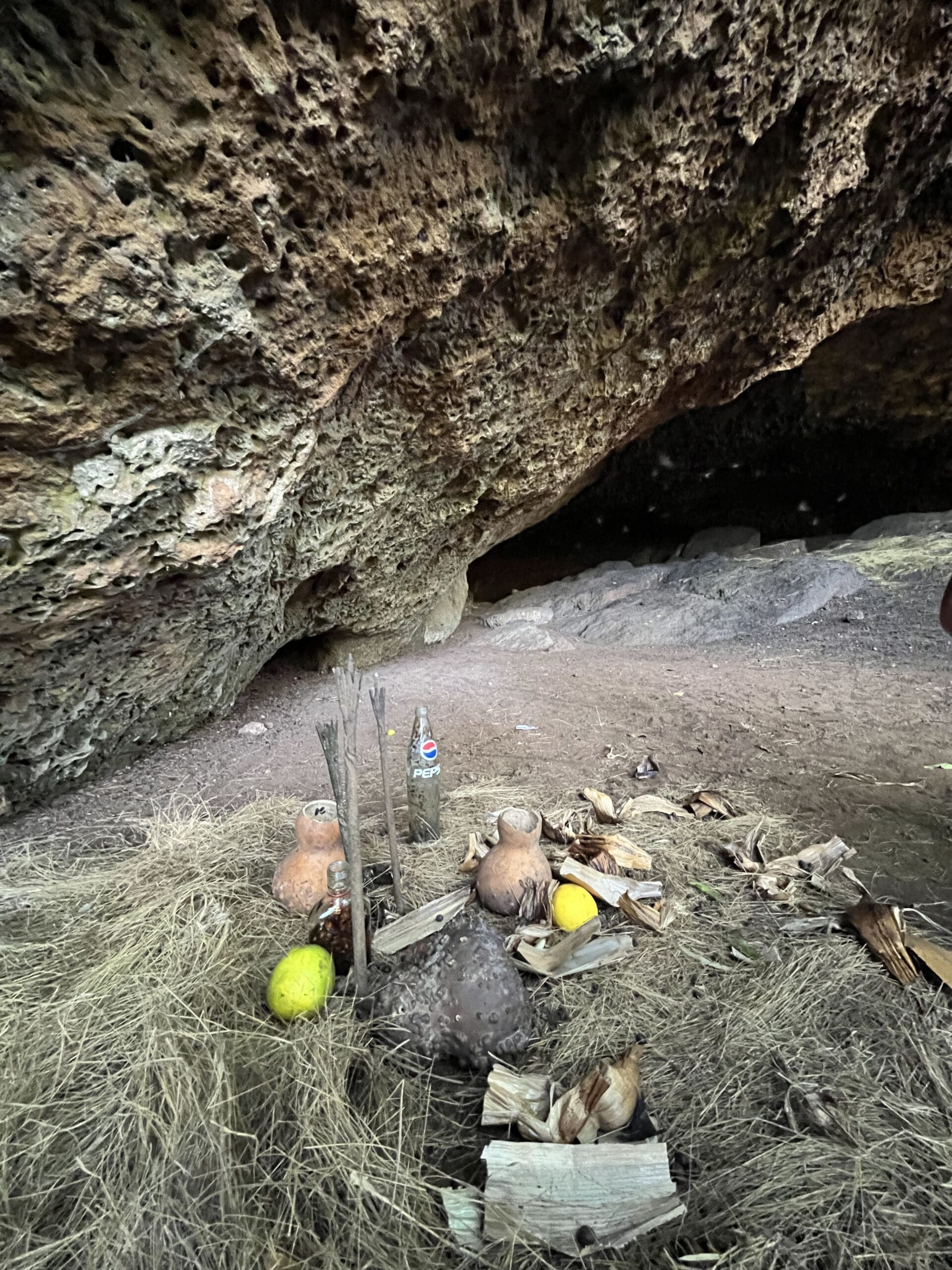 Een kleine grot met een ruw, rotsachtig plafond en droog gras op de grond. Bij de ingang liggen kalebassen, een citroen, een plastic fles en stokken die een offer of heiligdom vormen - misschien achtergelaten door reizigers die deze hoek van Afrika verkennen.
