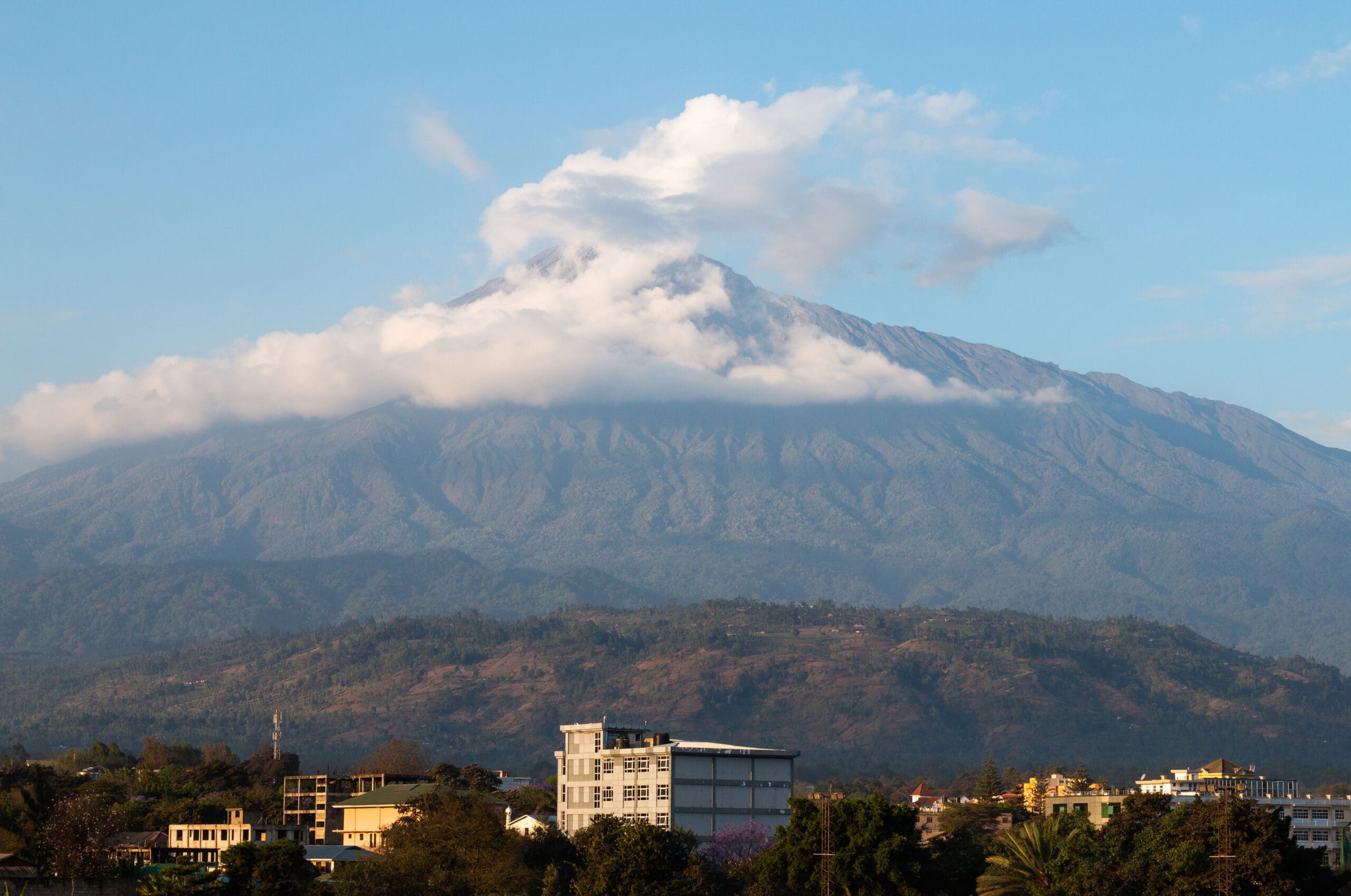 Een grote, met wolken bedekte berg rijst op boven een groen landschap en een Tanzaniaanse stad met gebouwen en bomen op de voorgrond onder een helderblauwe hemel, en biedt een verbluffende blik op Afrika voor avontuurlijke reizigers.