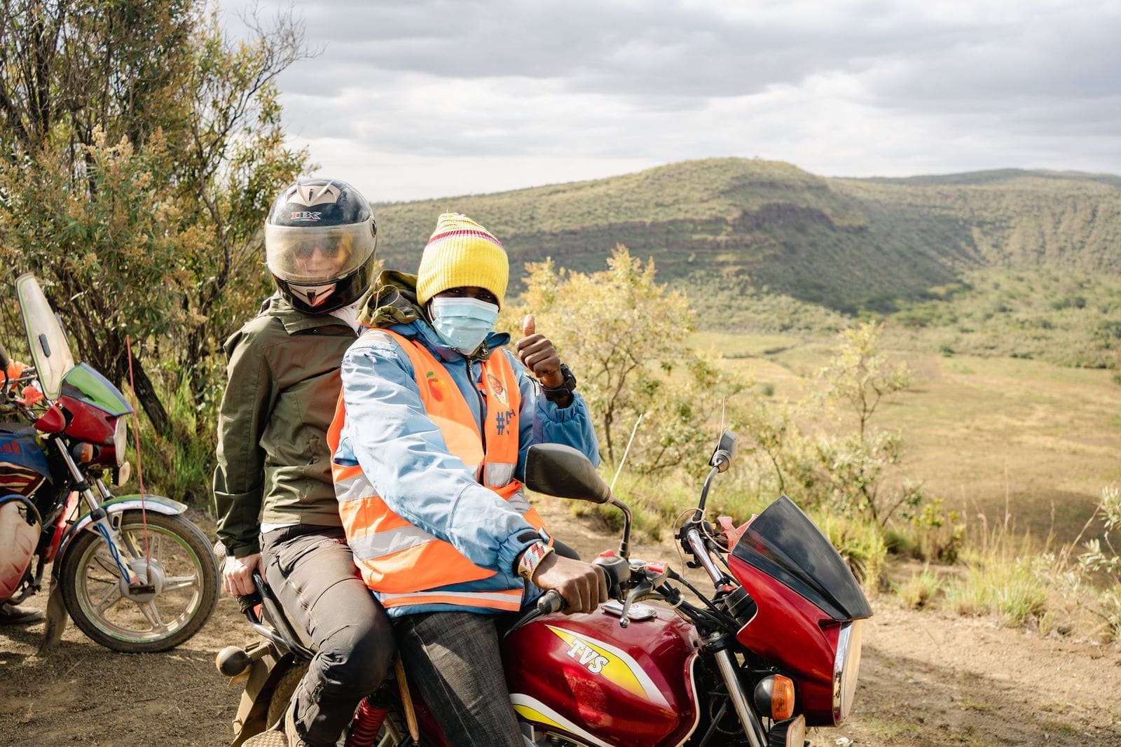 reiziger met helm achterop een boda boda motor met gids in fel oranje hesje in heuvelachtig landschap van kenia