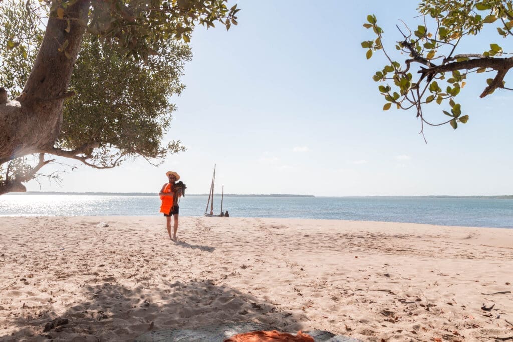 reiziger in fel oranje reddingsvest loopt over wit zandstrand met dhowboot op de achtergrond aan de kust van lamu kenia
