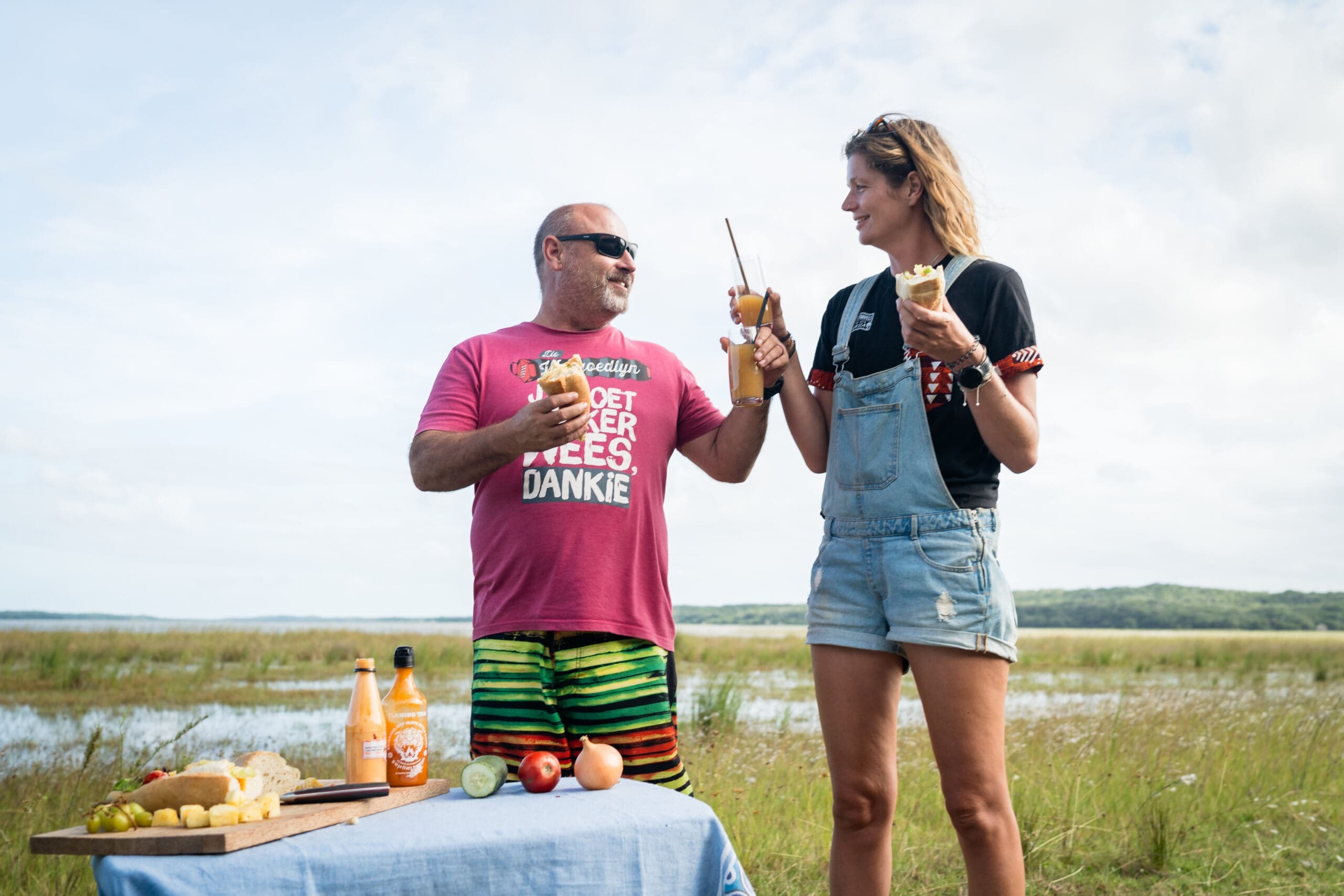 Een man en vrouw glimlachen en proosten met een drankje buiten bij een meer, genietend van een voorproefje van Mozambique. Voor hen staat een tafel met fruit en drankjes. De man draagt een rood shirt en een gestreepte korte broek; de vrouw draagt een overall en een zwart t-shirt.