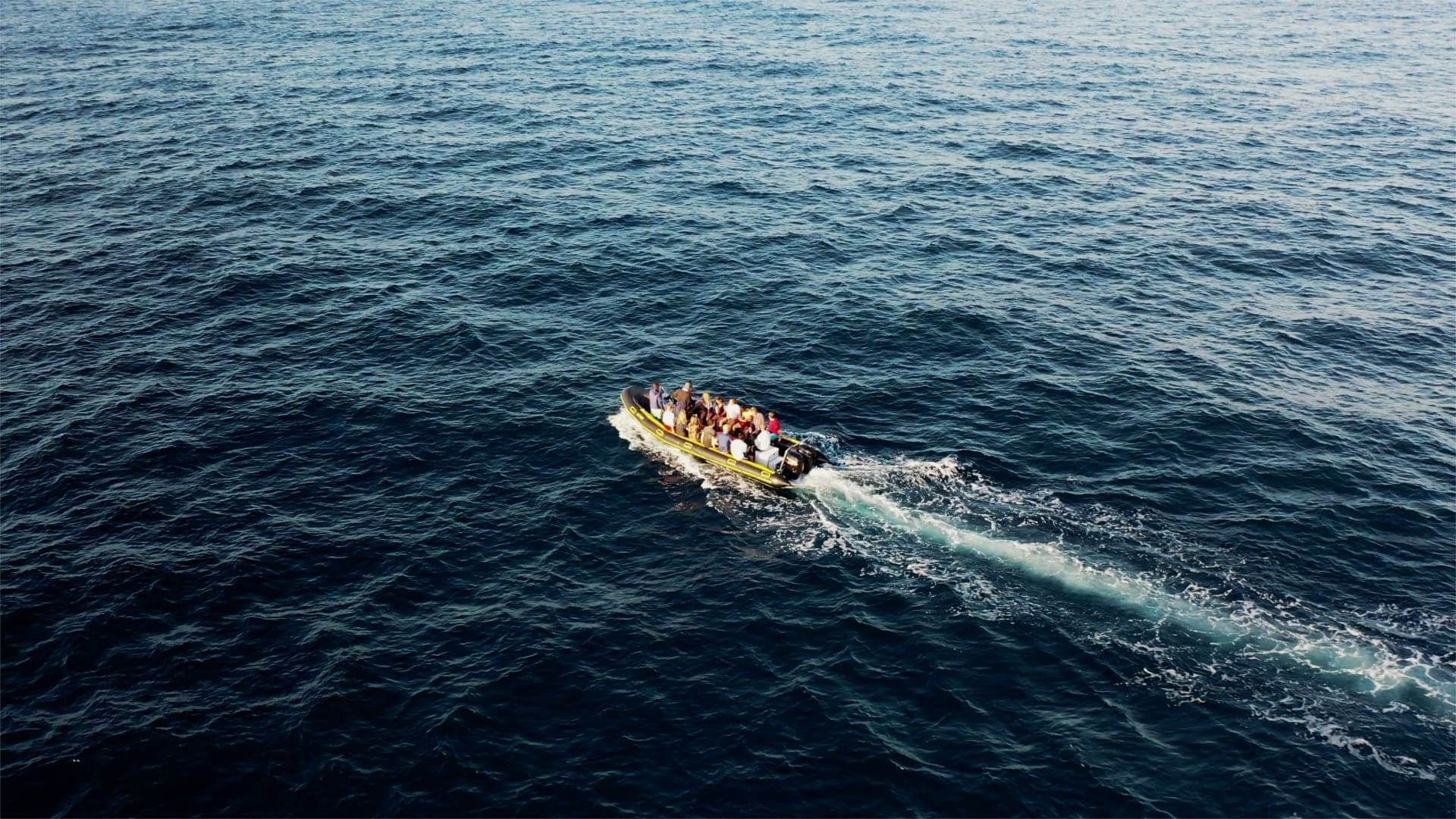Een kleine motorboot met meerdere mensen aan boord vaart over de uitgestrekte, kalme blauwe zee voor de kust van Mozambique en laat een lang wit kielzog achter zich. De boot lijkt weg te varen van de camera, omringd door open water in Afrika.