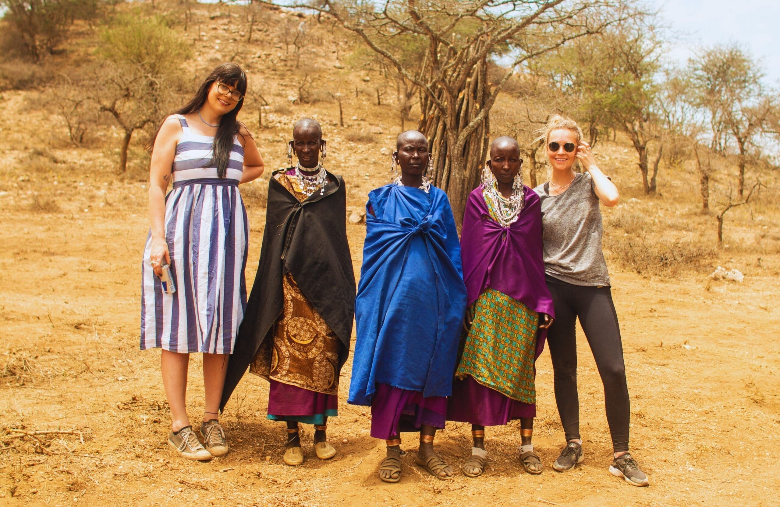 Twee vrouwen in vrijetijdskleding staan naast drie Maasai-vrouwen in traditionele kleding en sieraden, allemaal glimlachend buiten op een droge, aardachtige grond met bomen op de achtergrond - een gedenkwaardig moment van reizen door Tanzania in Afrika.