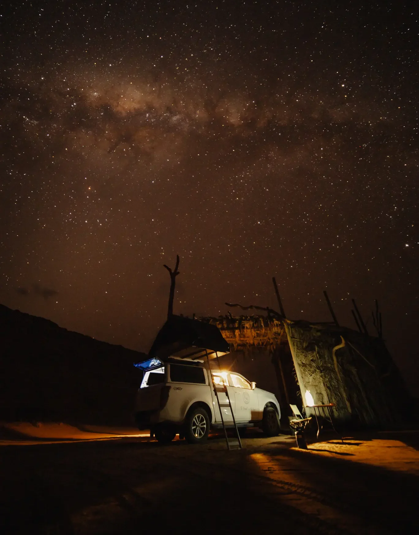 een verlichte terreinwagen met daktent staat geparkeerd bij een houten shelter in namibië onder een heldere sterrenhemel