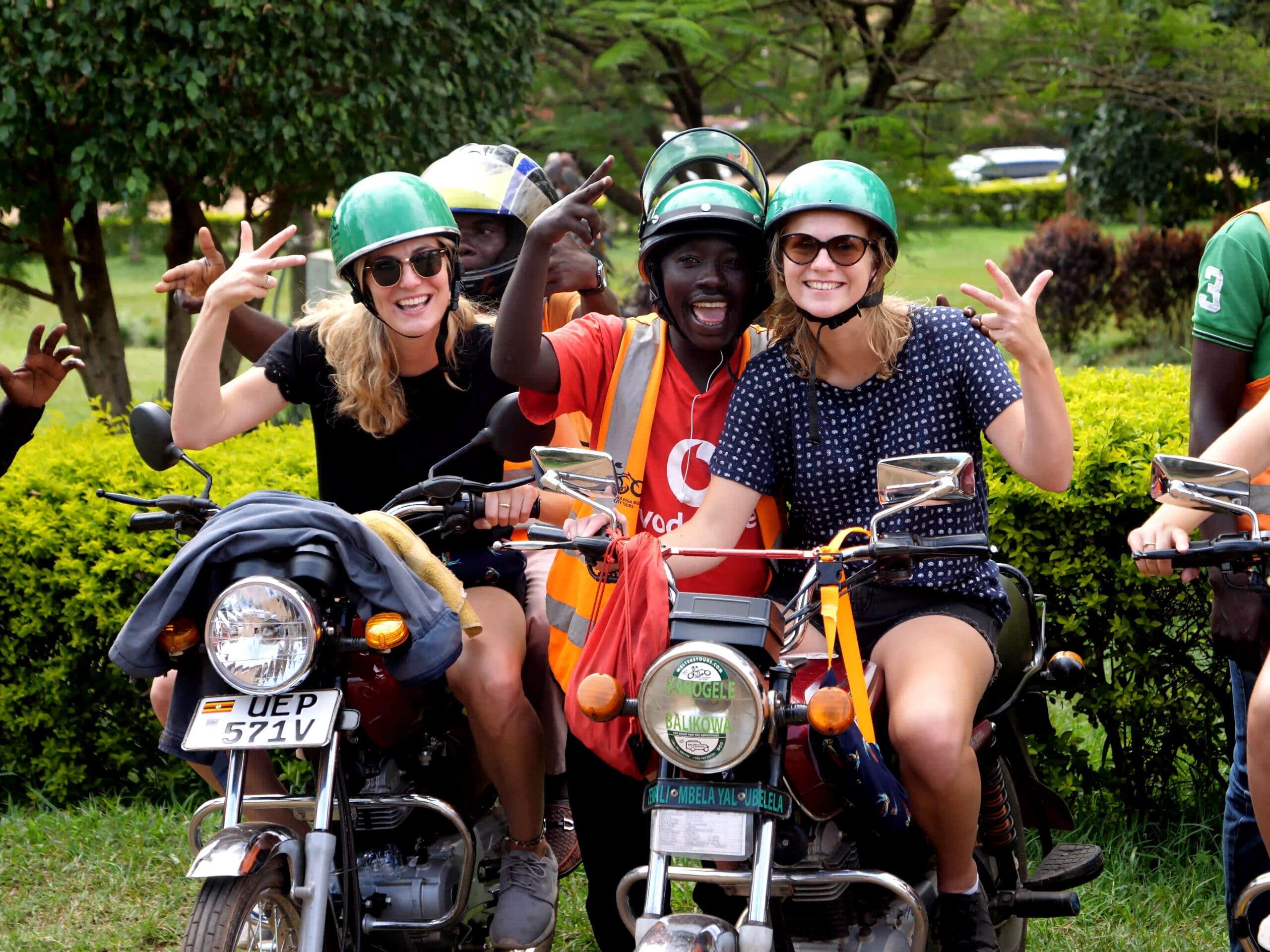 Drie lachende reizigers met helmen op zitten buiten op een motorfiets en poseren speels met vredestekens. Een persoon in een oranje vest staat tussen hen in, ook lachend en gebarend. Op de achtergrond zijn groen en bomen te zien.