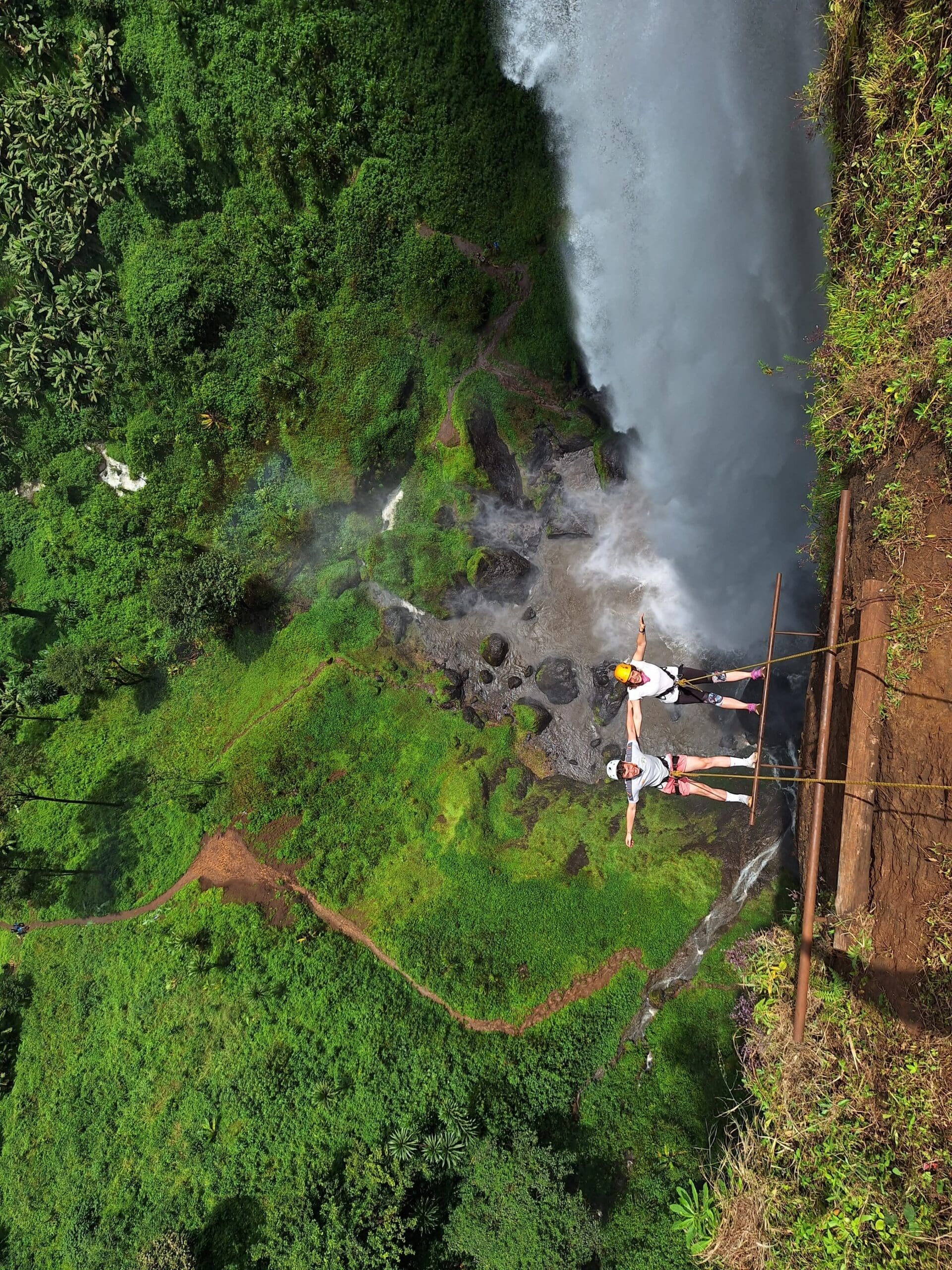 Twee reizigers staan op een houten platform, de armen wijd gespreid, naast een hoge waterval en een weelderig groene rotswand in Afrika, hoog boven de grond.