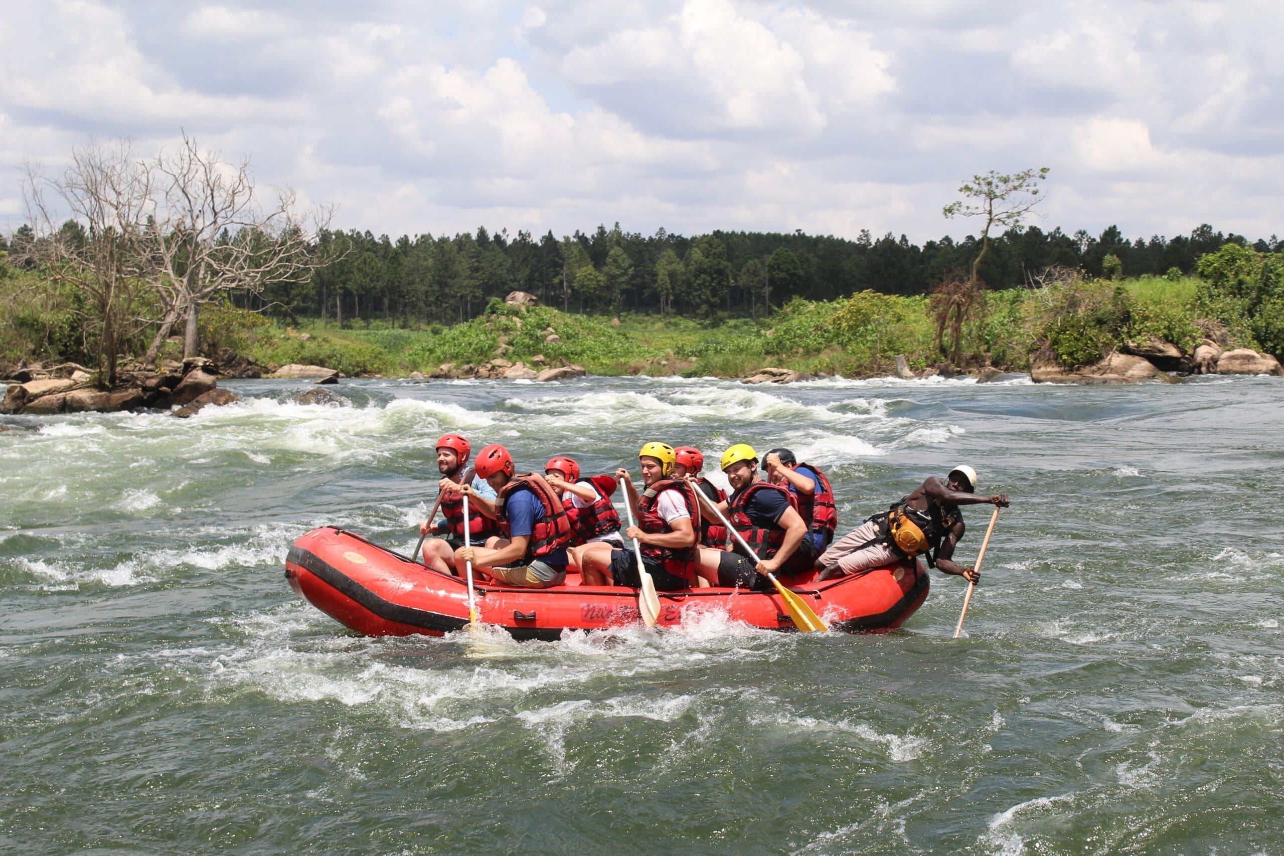Een groep reizigers met zwemvesten en helmen peddelt samen in een rood opblaasbaar vlot op een rivier met stroomversnellingen, omringd door groene bomen en een halfbewolkte hemel - een onvergetelijk reizigersavontuur.