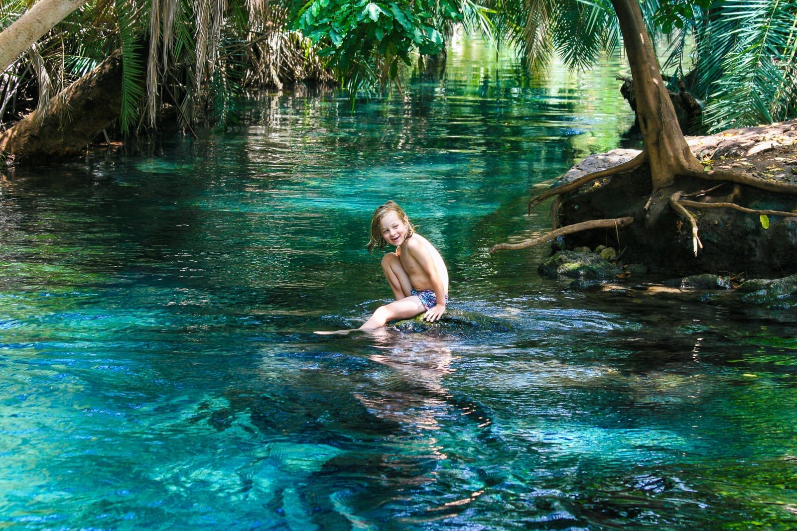Een jong kind in zwemkleding zit en lacht op een rots in een helder, blauw natuurlijk zwembad omringd door weelderige groene bomen en planten - een beeld van de schoonheid van reizen door de prachtige landschappen van Afrika.