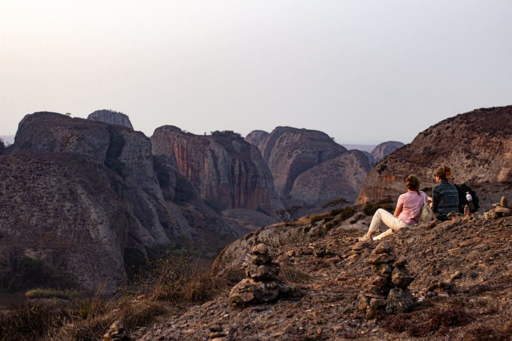 Two people sit on a rocky hillside in Angola, surrounded by stacked stone piles. They overlook large, rounded rock formations and a wide landscape under a hazy sky—a perfect moment for travelling in Africa.
