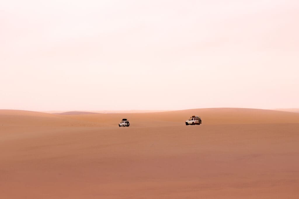 Twee witte terreinwagens rijden door een uitgestrekte, lege woestijn in Angola met gladde, zanderige duinen onder een bleke, pastelkleurige hemel. Het tafereel voelt afgelegen en sereen aan - perfect voor avontuurlijke reizigers die op zoek zijn naar de ongerepte schoonheid van Afrika.