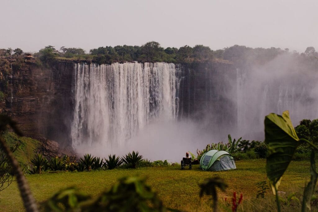 A green tent and camping chairs are set up on grassy land in front of a large, misty waterfall with lush greenery in the background, capturing the spirit of travelling through Angola’s breathtaking landscapes.