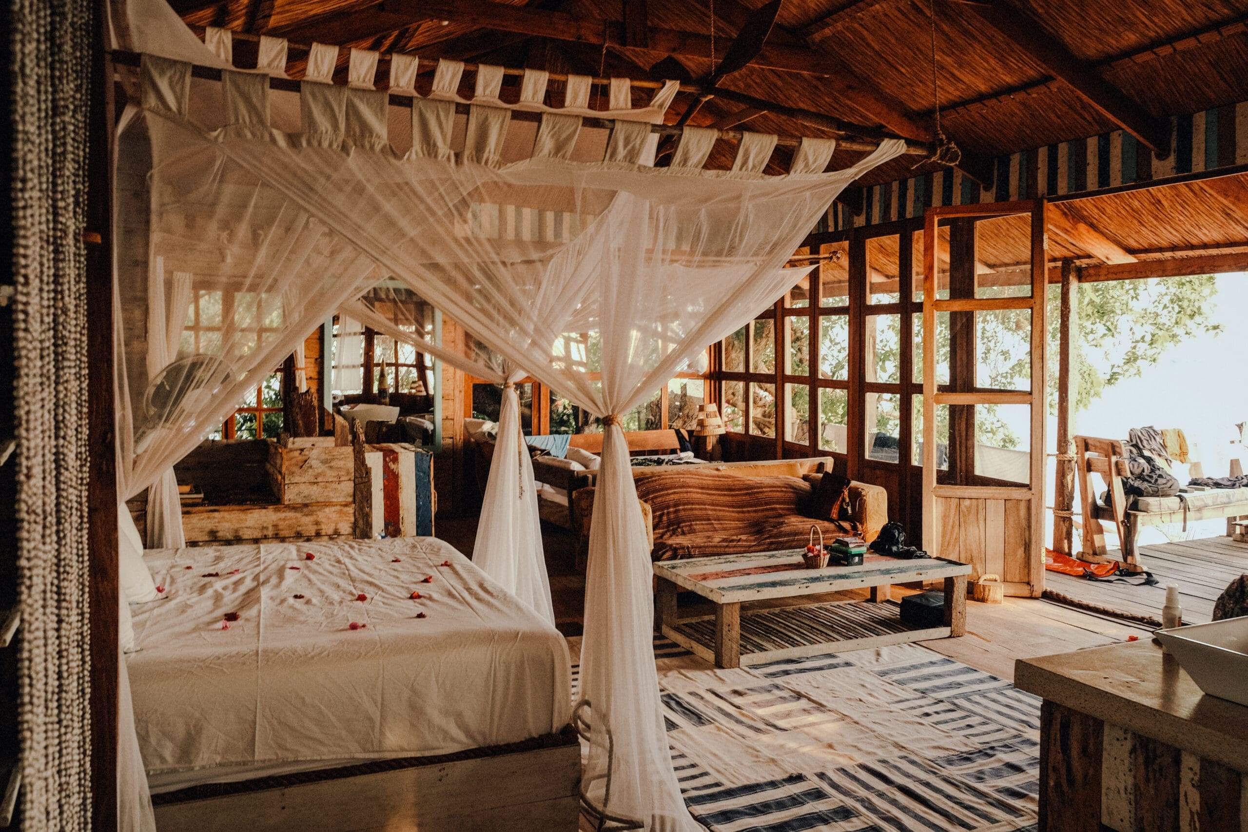 Een gezellige, rustieke slaapkamer in Malawi met een hemelbed gedrapeerd in wit muskietengaas, houten muren en plafond, gestreepte tapijten, een bank en zonlicht dat door de grote ramen naar binnen schijnt - perfect voor Afrika reizen.