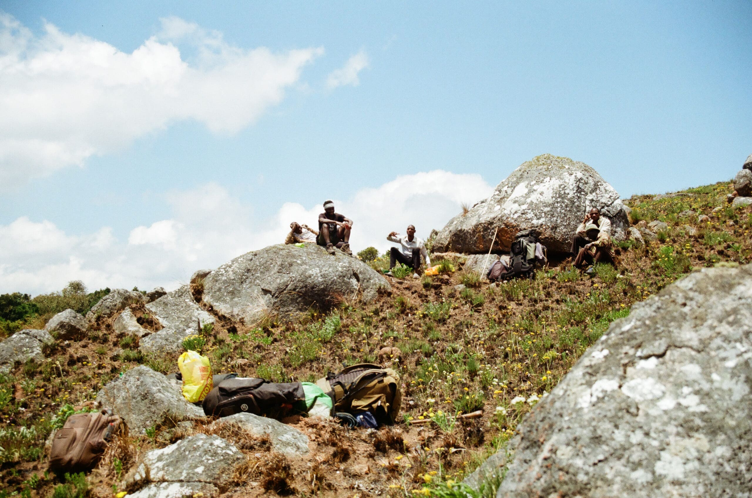 Verschillende reizigers zitten en rusten op grote rotsen op een met gras begroeide, hellende heuvel onder een gedeeltelijk bewolkte hemel. Rugzakken en spullen liggen verspreid in de buurt, wat duidt op een pauze tijdens hun trektocht door Malawi.