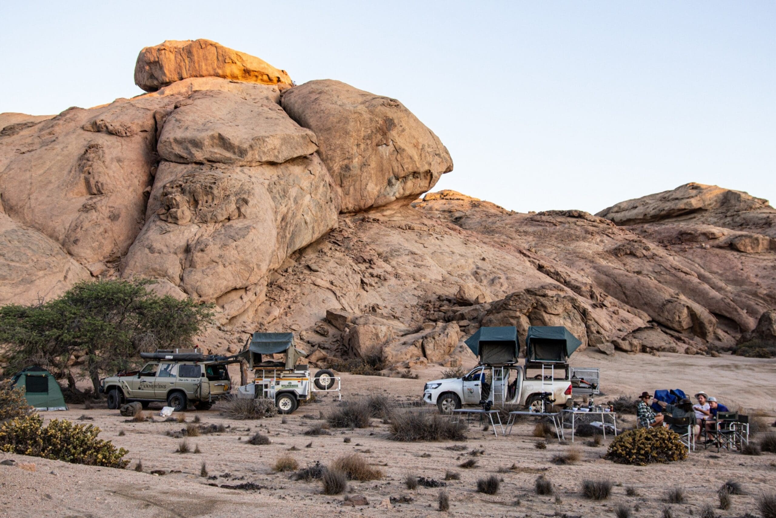 Twee terreinwagens met daktenten staan geparkeerd bij rotsblokken in een woestijnlandschap. Kampeerspullen en tafels staan vlakbij opgesteld, met een paar mensen eromheen. Op de voorgrond is schaarse vegetatie te zien.