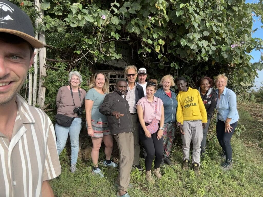 Een groep mensen, zowel mannen als vrouwen, staan dicht bij elkaar buiten voor weelderig groen gebladerte en glimlachen voor de camera. Een man met een pet staat op de voorgrond en neemt de groepsselfie.