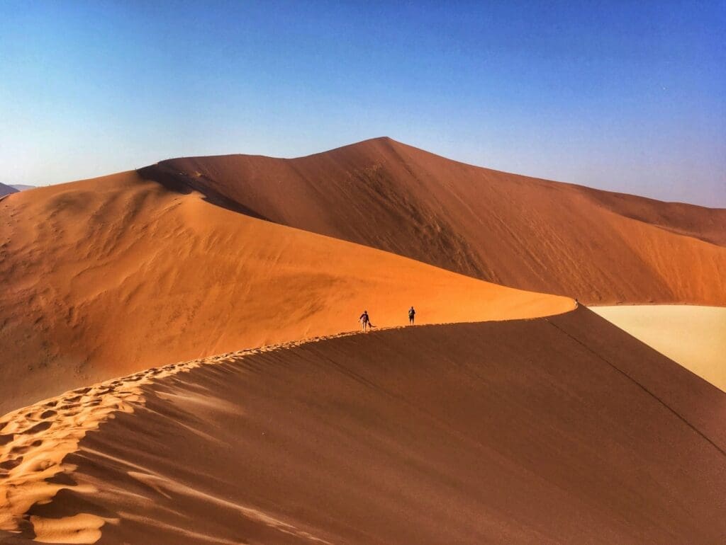 Zwei Personen spazieren auf dem Kamm einer großen Sanddüne unter blauem Himmel, umgeben von weiten, sonnenbeschienenen Wüstendünen.
