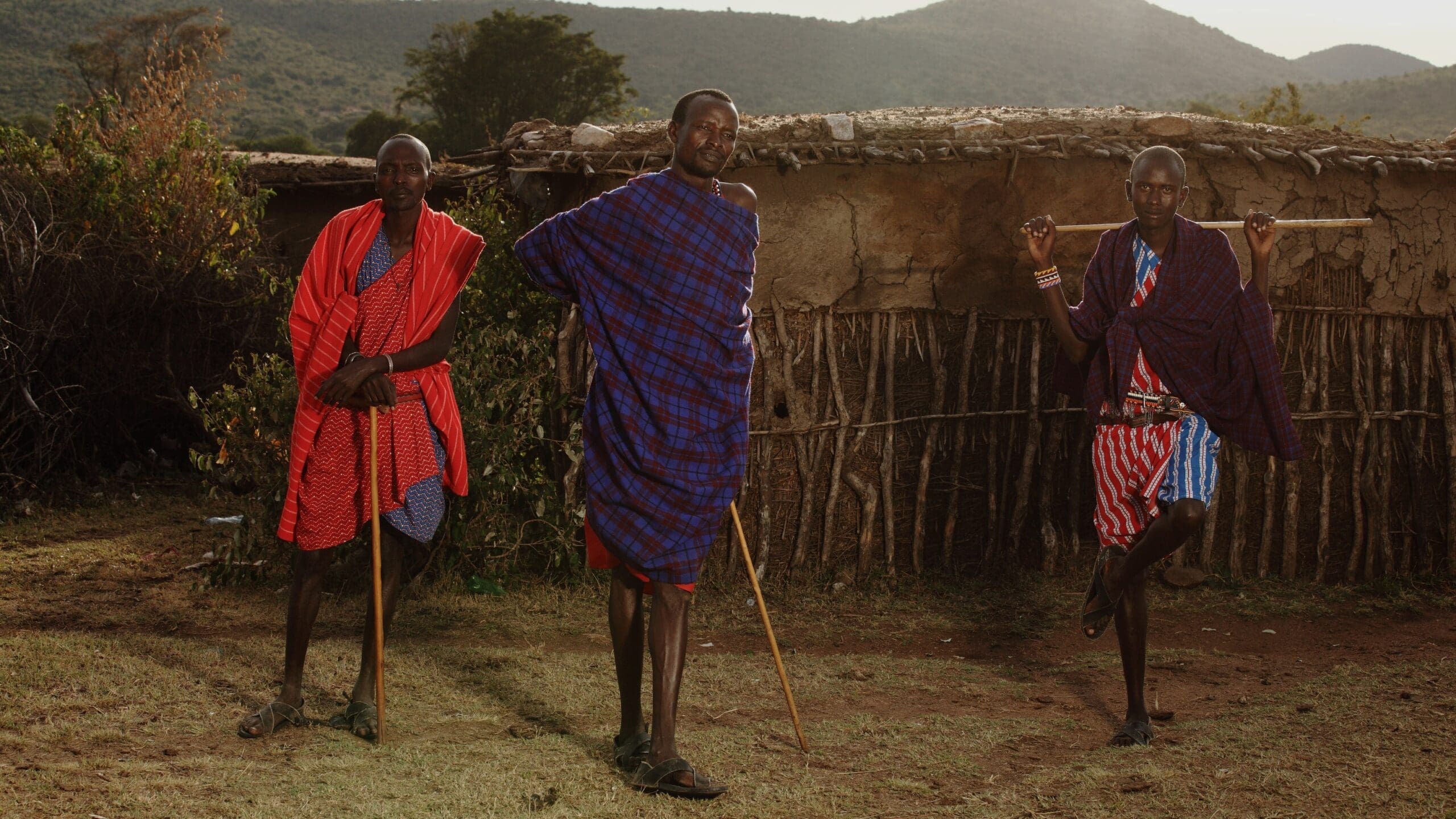 Drie Maasai mannen staan buiten in traditionele shuka's met wandelstokken voor een rieten hut met bergen op de achtergrond. De mannen poseren zelfverzekerd op een grasveld onder warm zonlicht.