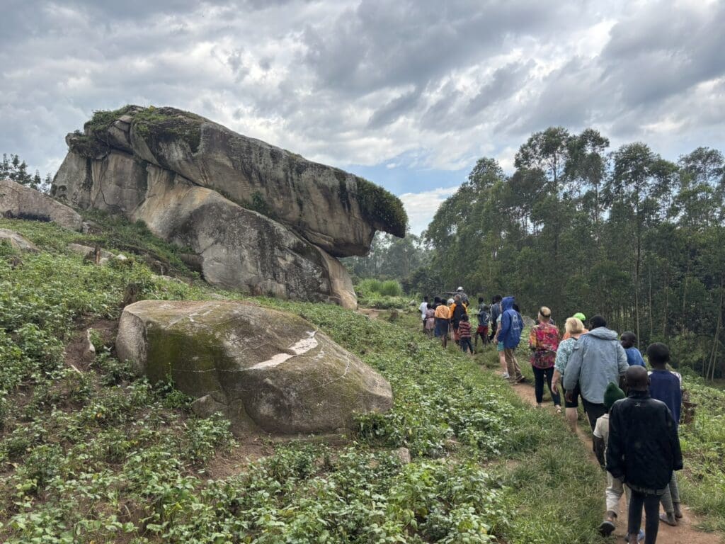 Een groep mensen loopt over een onverhard pad door groene vegetatie naar een grote, overhangende rotsformatie onder een bewolkte hemel, met hoge bomen op de achtergrond.