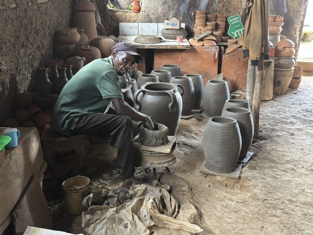 Een man zit in een werkplaats klei te vormen op een pottenbakkersschijf, omringd door voltooide potten en vazen. Planken en tafels tonen meer aardewerk en gereedschap in de rustieke, aardse ruimte.
