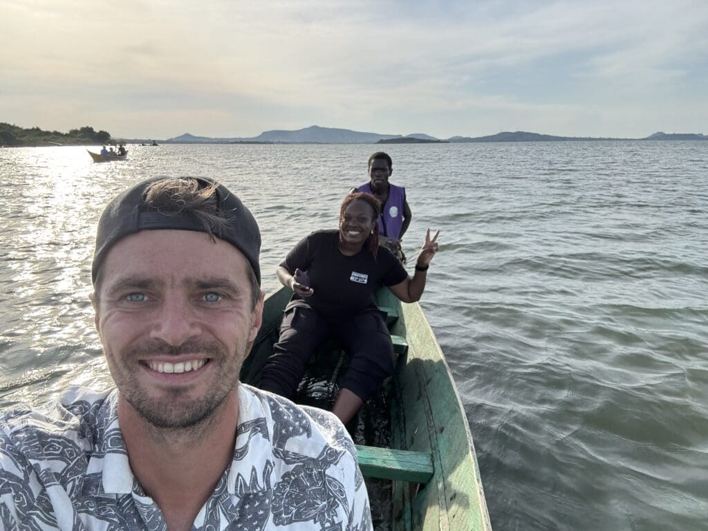 Drie mensen glimlachen terwijl ze in een kleine houten boot op een meer zitten met heuvels in de verte en een gedeeltelijk bewolkte lucht op de achtergrond. De voorste persoon neemt een selfie en één persoon maakt een vredesteken.