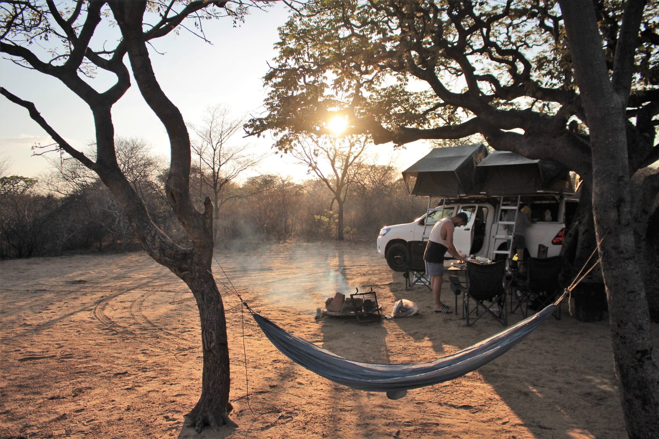Een camping bij zonsondergang met een witte SUV, daktenten, een kampvuur, campingstoelen en een hangmat tussen twee bomen. Een persoon staat bij het vuur te midden van droge bomen en zanderige grond.