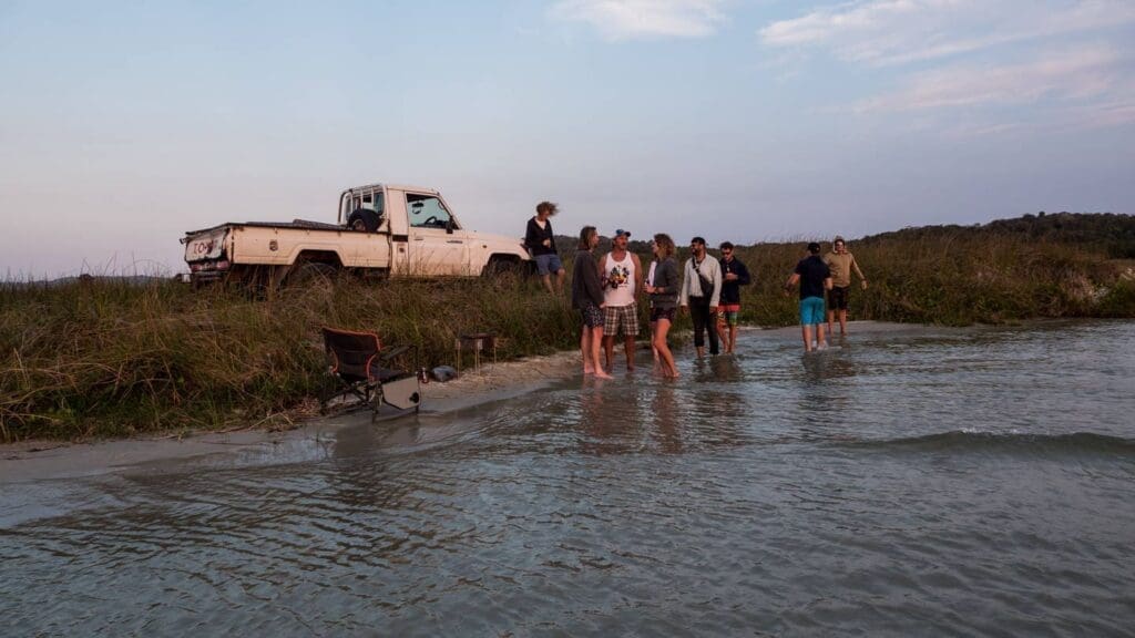 Een groep mensen verzamelt zich aan de rand van een meer of rivier bij een met gras begroeide oever, met een geparkeerde witte pick-up truck en kampeerspullen in de buurt tijdens zonsondergang. Sommige mensen staan in het ondiepe water, anderen op de oever.