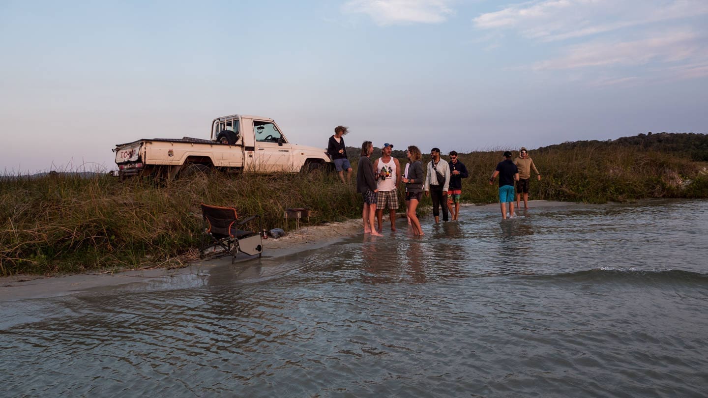 Een groep mensen verzamelt zich aan de rand van een meer of rivier bij een met gras begroeide oever, met een geparkeerde witte pick-up truck en kampeerspullen in de buurt tijdens zonsondergang. Sommige mensen staan in het ondiepe water, anderen op de oever.