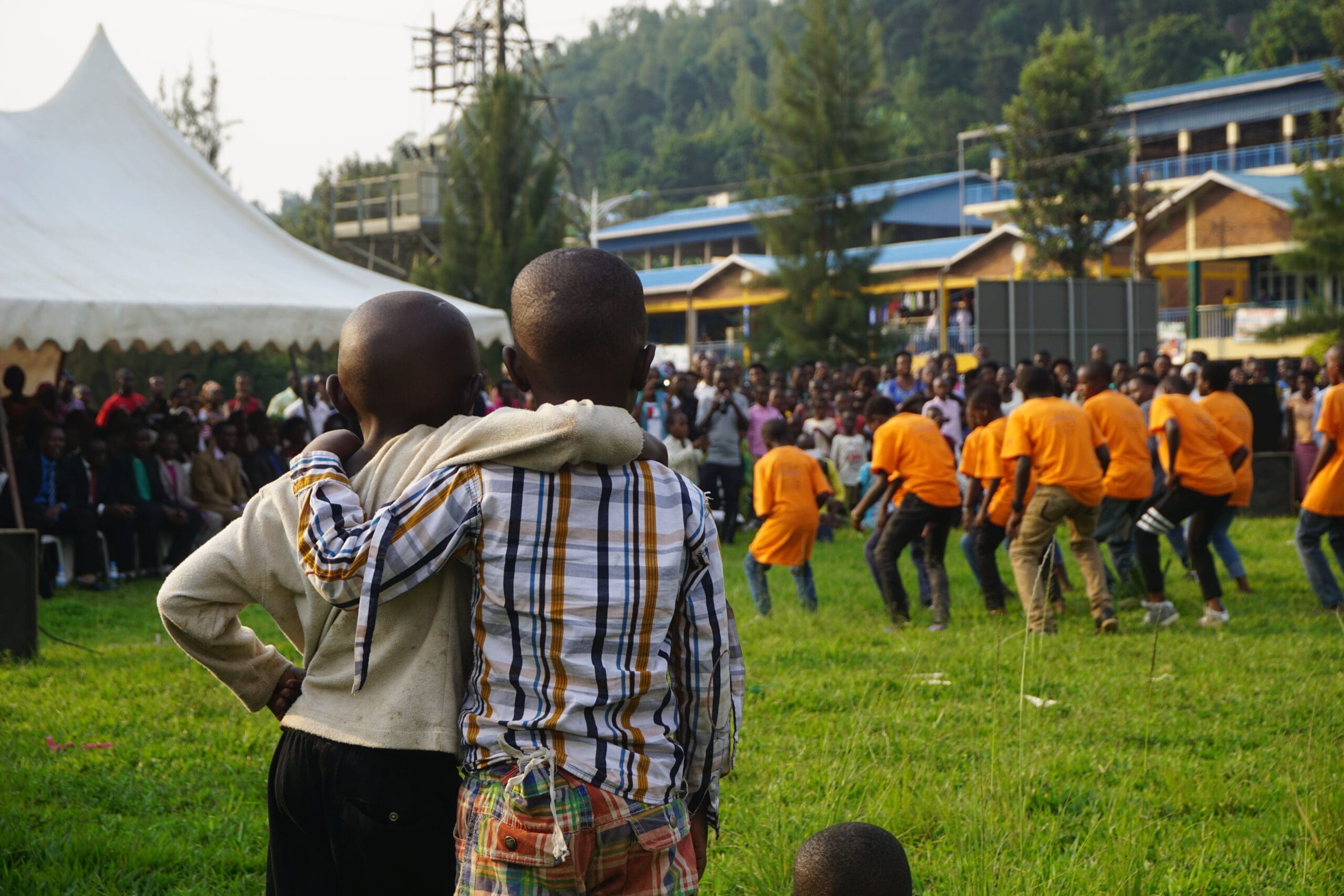 Twee kinderen staan op het gras met hun armen om elkaar heen en kijken naar een groep mensen in oranje shirts die buiten optreden. Een grote menigte zit onder een witte tent op de achtergrond, met bomen en gebouwen zichtbaar.