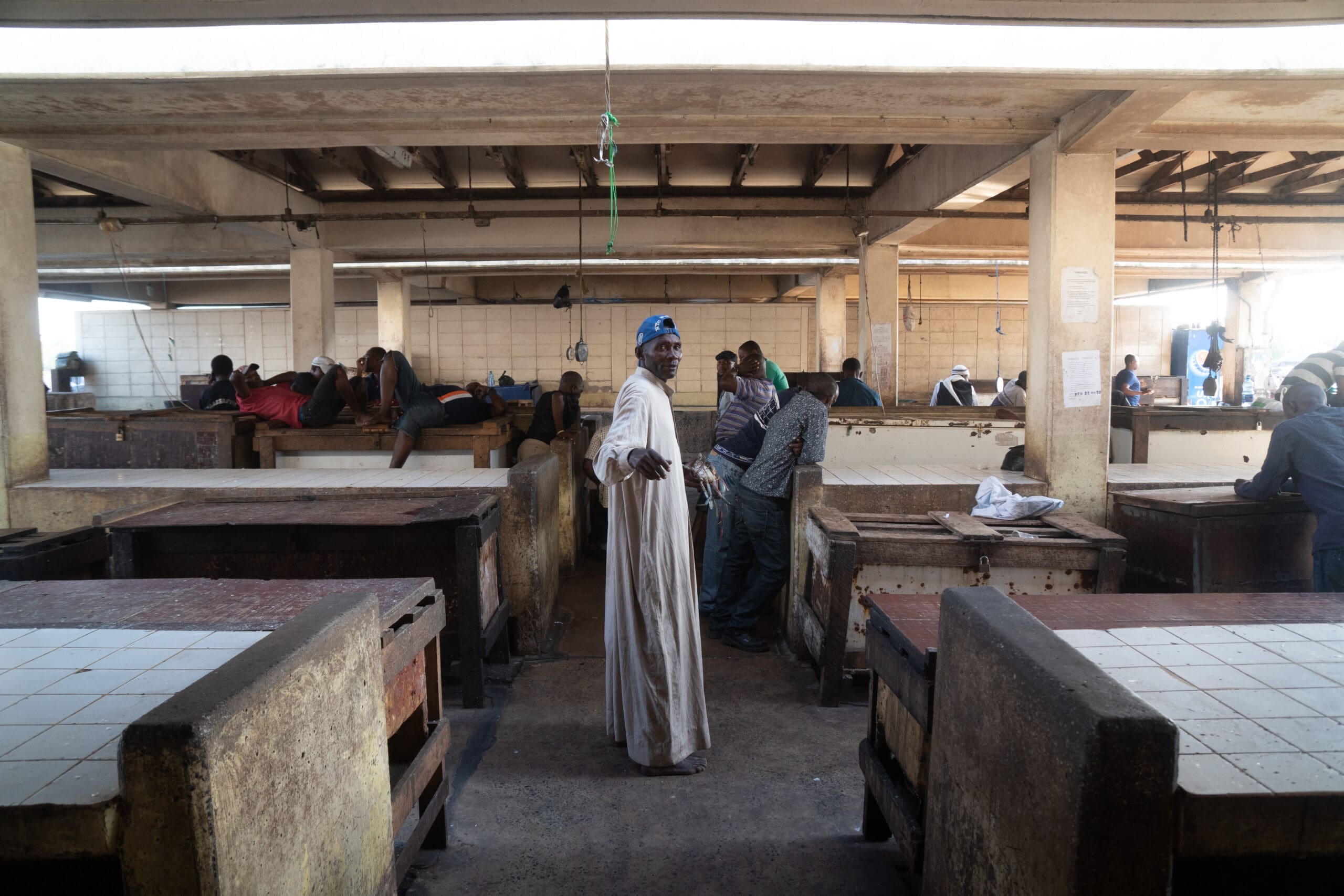 Een persoon in een lang wit gewaad en blauwe hoofddoek staat midden op een overdekte markt, kijkt achterom en glimlacht. Andere mensen zitten of staan eromheen, met lege marktkramen op de voorgrond.