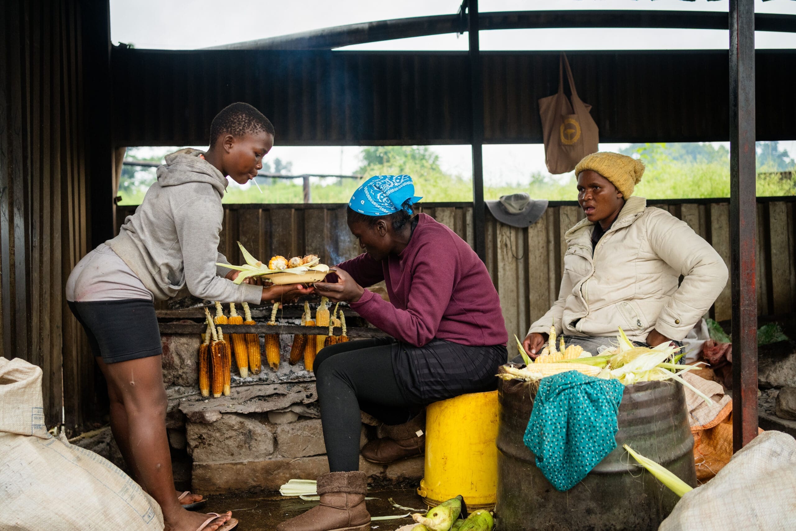 Drie vrouwen bereiden en grillen maïskolven in een buitenruimte. Twee vrouwen bereiden maïs boven een open grill, terwijl een derde vrouw, rechts zittend, toekijkt. Om hen heen liggen maïskolven en zakken verspreid.