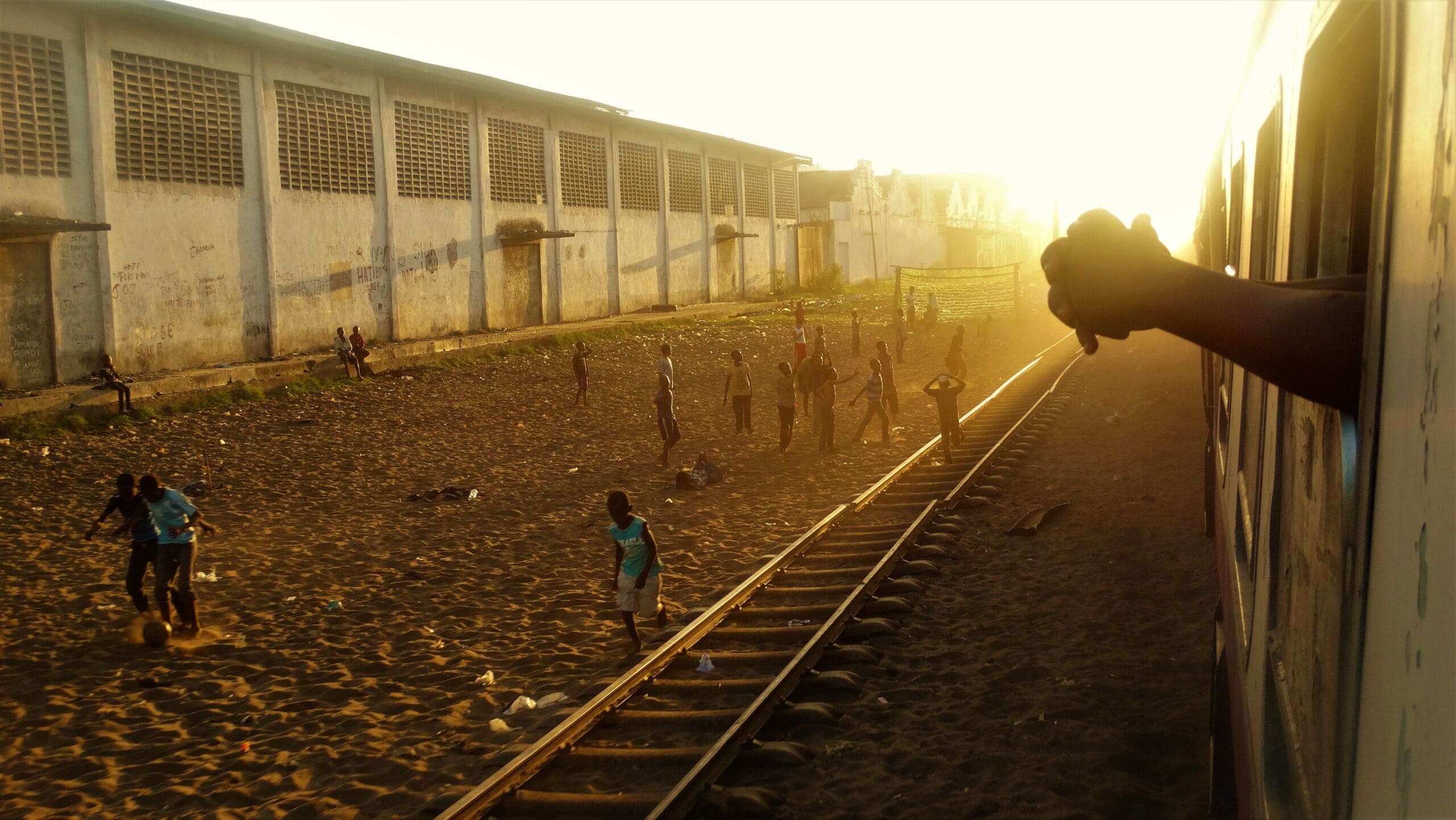 Kinderen voetballen op een zanderig veld naast treinrails bij zonsondergang, vlakbij een groot, verweerd gebouw, terwijl iemands hand uitsteekt naar een passerende trein op de voorgrond.