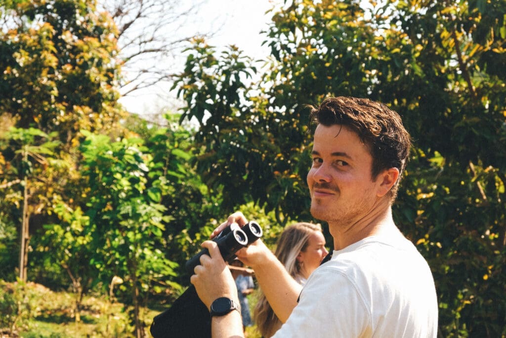 Een man glimlacht naar de camera terwijl hij een verrekijker vasthoudt in een weelderige, zonovergoten tuin. Een andere persoon staat op de achtergrond, gedeeltelijk zichtbaar tussen groene bomen en planten.