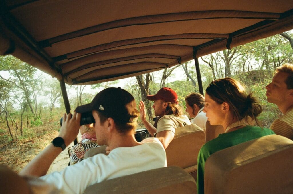 Een groep mensen zit in een safarivoertuig dat door een bebost gebied rijdt. Eén persoon gebruikt een verrekijker, terwijl anderen vooruit kijken, sommigen met hoeden en zonnebrillen op. Zonlicht filtert door de bomen.