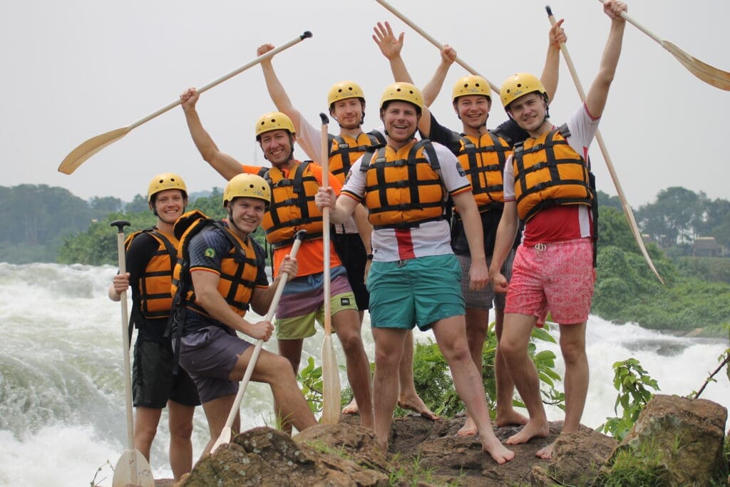 Een groep van acht mensen in zwemvesten en helmen poseert vrolijk met peddels op een rotsachtige rivieroever, met wildwaterbaan en groen op de achtergrond.
