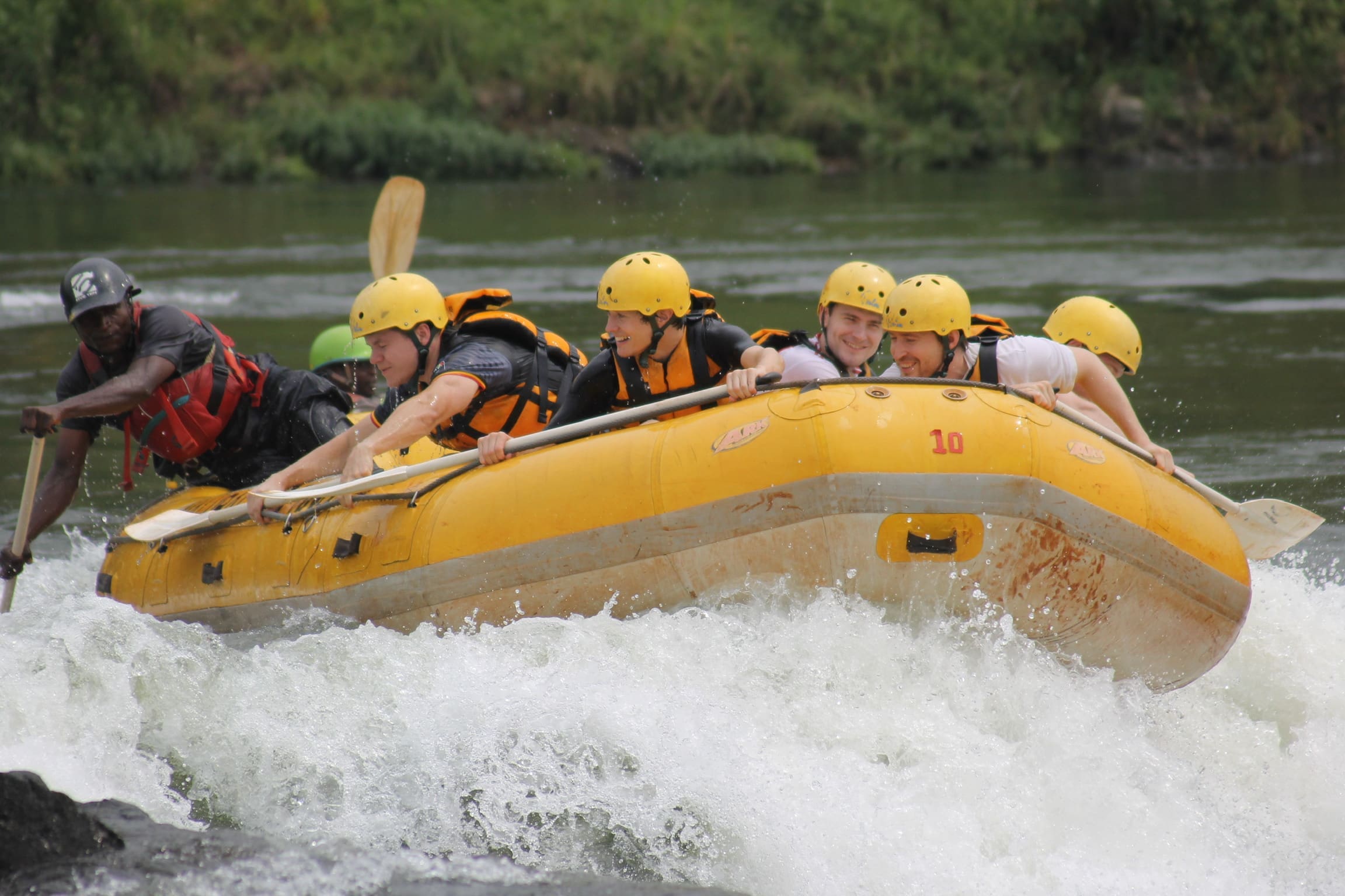Zes mensen met gele helmen en reddingsvesten varen op een geel opblaasbaar vlot door woelig water met een gids voorop. Groene vegetatie omzoomt de rivieroever op de achtergrond.