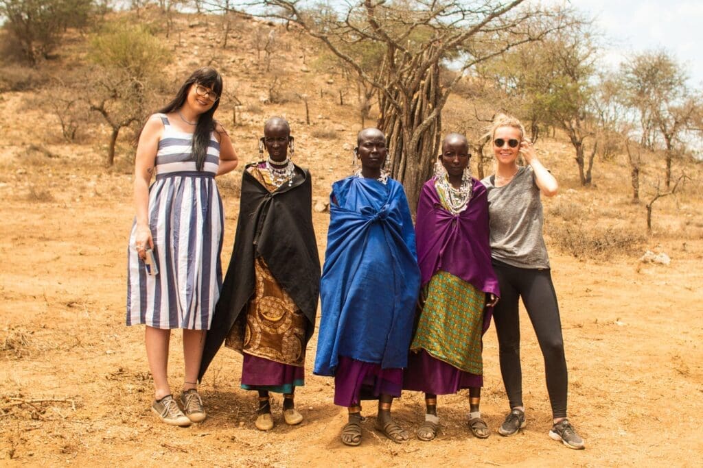 Twee vrouwen staan met drie Maasai-vrouwen gekleed in traditionele kleding en sieraden en poseren samen buiten op een droog, stoffig land met schaarse bomen en heuvels op de achtergrond.