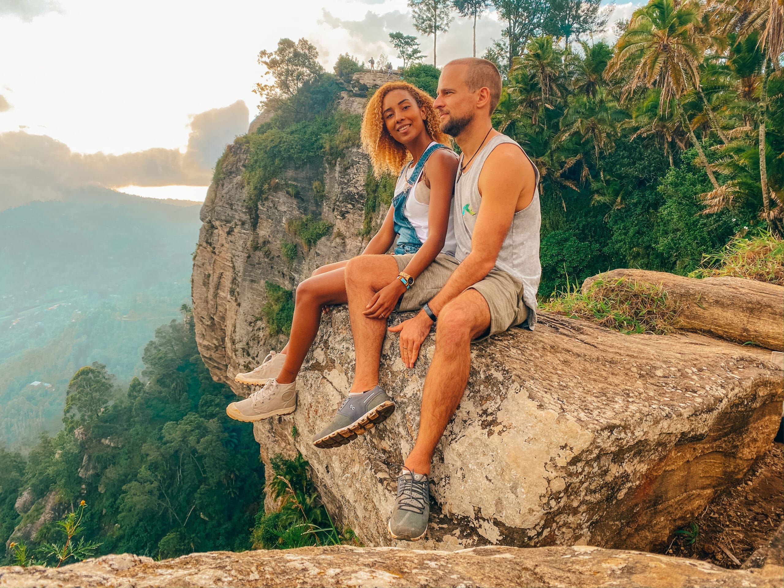 Een man en vrouw zitten samen op een grote rotsrichel met uitzicht op een weelderige, groene vallei met hoge bomen en bergen in de verte onder een bewolkte hemel. Ze lijken ontspannen en genieten van het schilderachtige uitzicht.