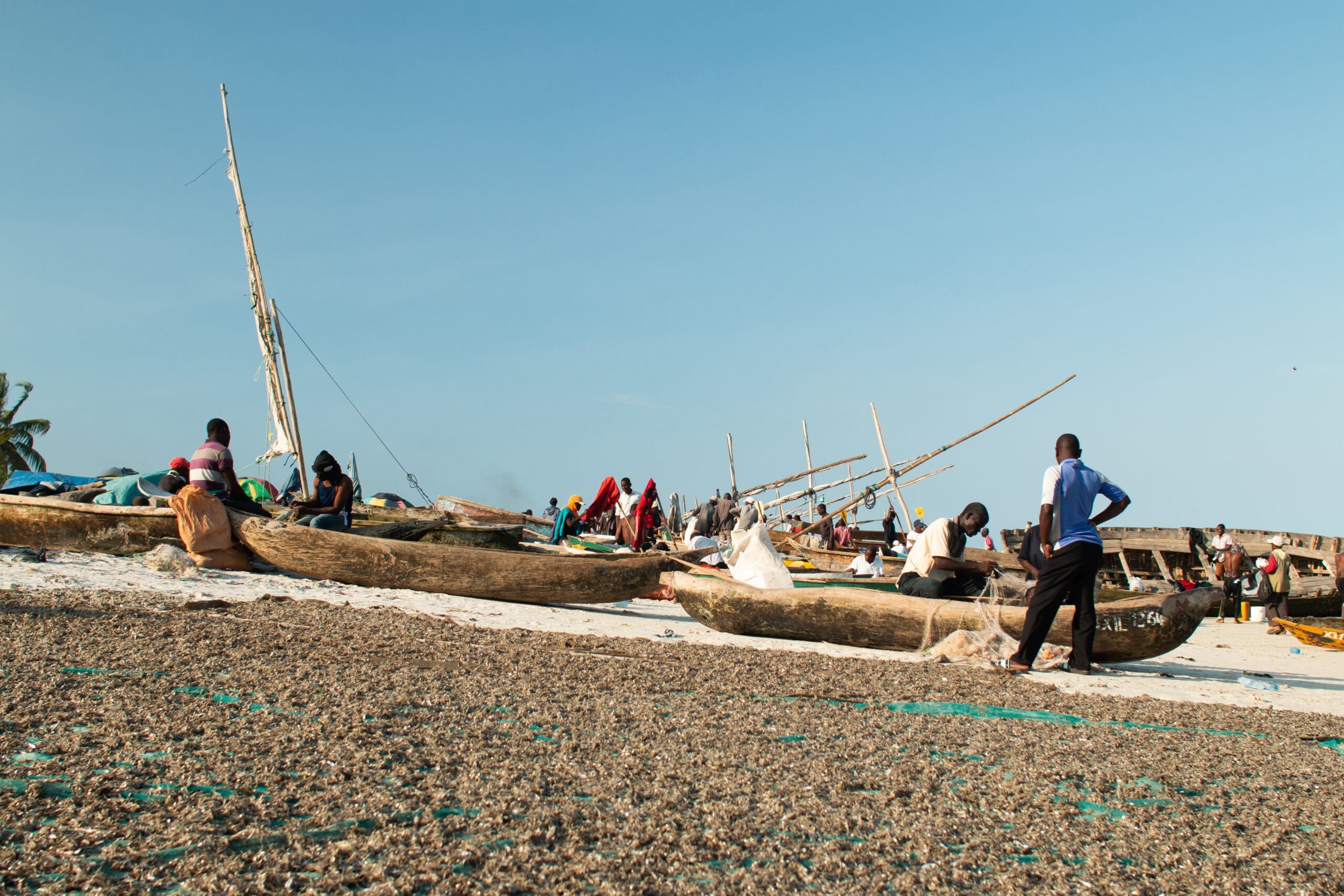 Mensen verzamelen zich rond houten vissersboten op een zandstrand onder een strakblauwe hemel. Sommigen zitten op de boten terwijl anderen in de buurt staan of lopen. Netten en vistuig liggen verspreid over de kust.