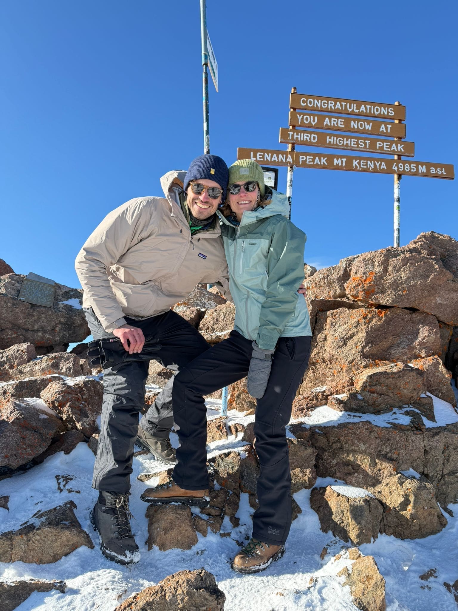 Twee mensen in winterjassen en laarzen glimlachen en poseren op de besneeuwde, rotsachtige grond op Lenana Peak, Mt. Kenya, met felicitatieborden voor het bereiken van de op twee na hoogste piek op de achtergrond onder een strakblauwe hemel.