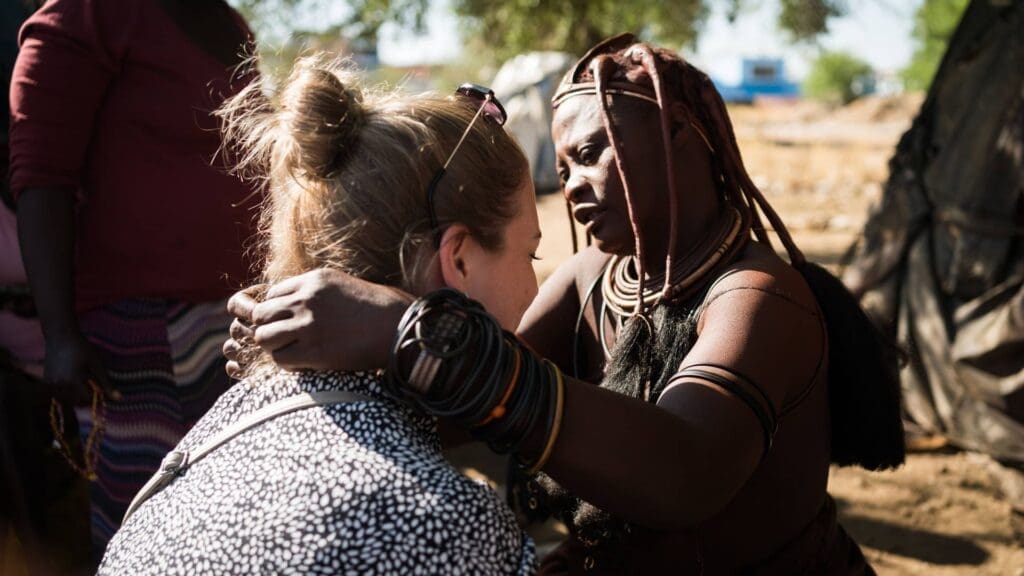 Een Himba vrouw met traditionele juwelen en kleding raakt zachtjes de schouders aan van een zittende vrouw met een lichte huid en blond haar, die van de camera weg kijkt, in een buitenopstelling.