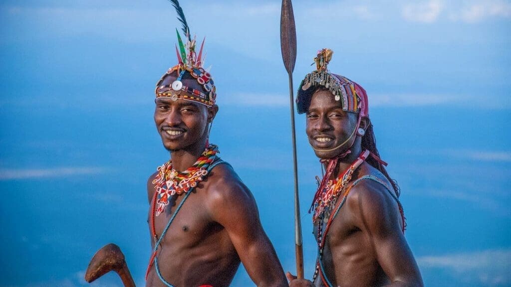Twee Maasai mannen staan naast elkaar en glimlachen, dragen traditionele kralen sieraden en hoofddeksels, de een houdt een speer vast en de ander een houten knots, met een uitgestrekte blauwe lucht op de achtergrond.