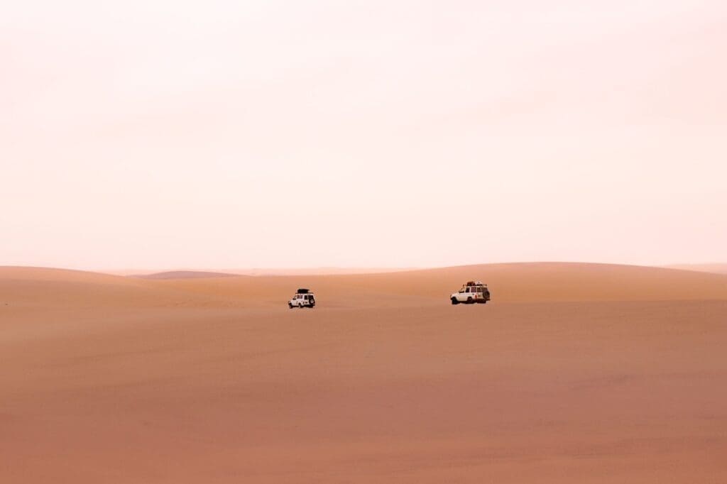 Zwei weiße Geländewagen fahren durch eine weite, leere Wüstenlandschaft unter einem blassen, dunstigen Himmel. Die Sanddünen sind glatt und weitläufig, mit weichen, warmen Farbtönen. Die Szene wirkt ruhig und isoliert.