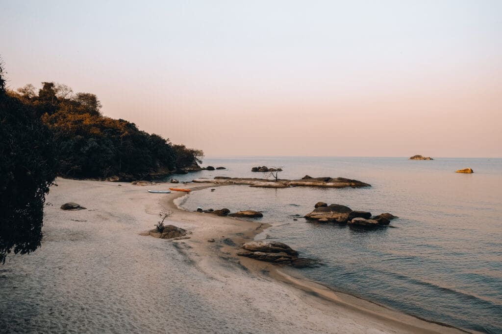 Een rustig strandtafereel bij zonsondergang met kalm water, grote rotsen verspreid langs de kust, zandstrand, kajaks op het zand en bomen langs de kustlijn onder een zachte, roze lucht.
