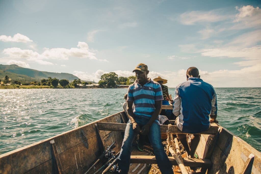 Drei Männer sitzen in einem Holzboot auf einem See unter einem teilweise bewölkten Himmel, mit grünen Hügeln und einer Uferlinie im Hintergrund. Das Wasser ist ruhig und das Sonnenlicht erhellt die Szene.