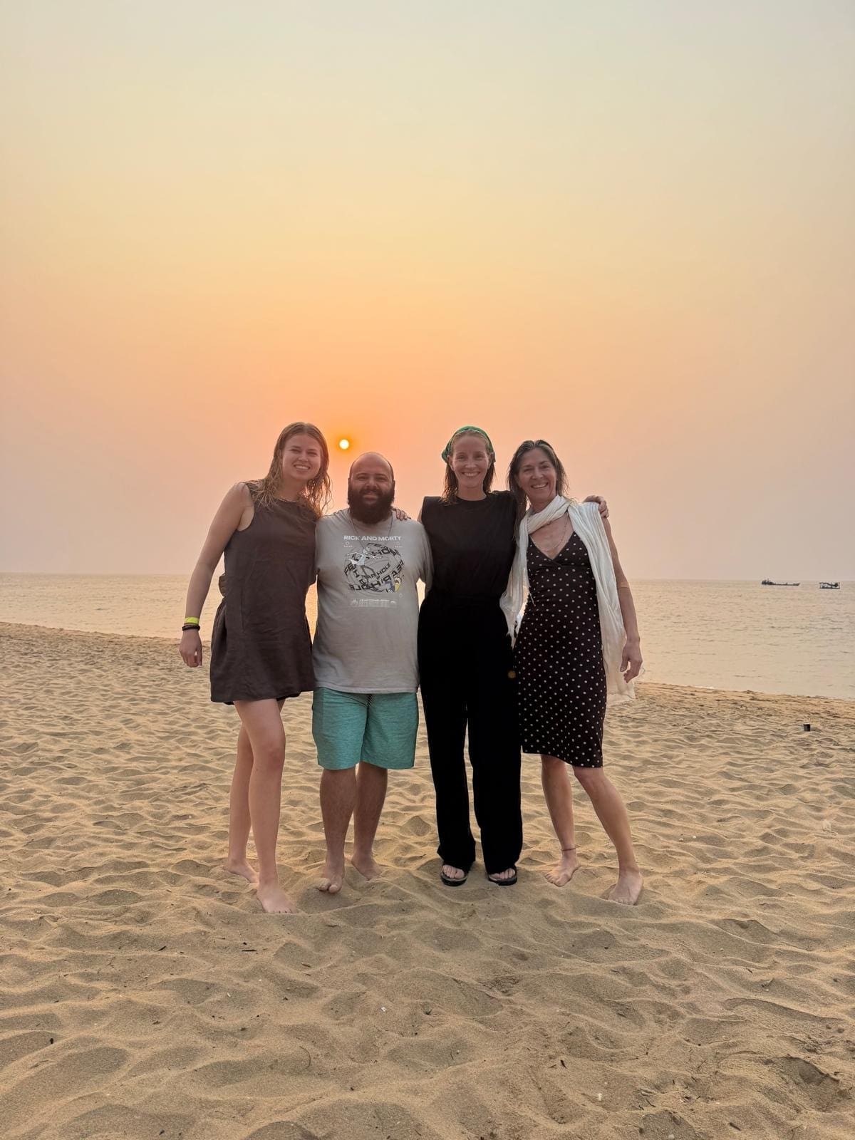 Vier mensen staan op blote voeten op een zandstrand bij zonsondergang, glimlachen en poseren samen met de zee en een oranje zon op de achtergrond. De lucht is helder en zacht verlicht.