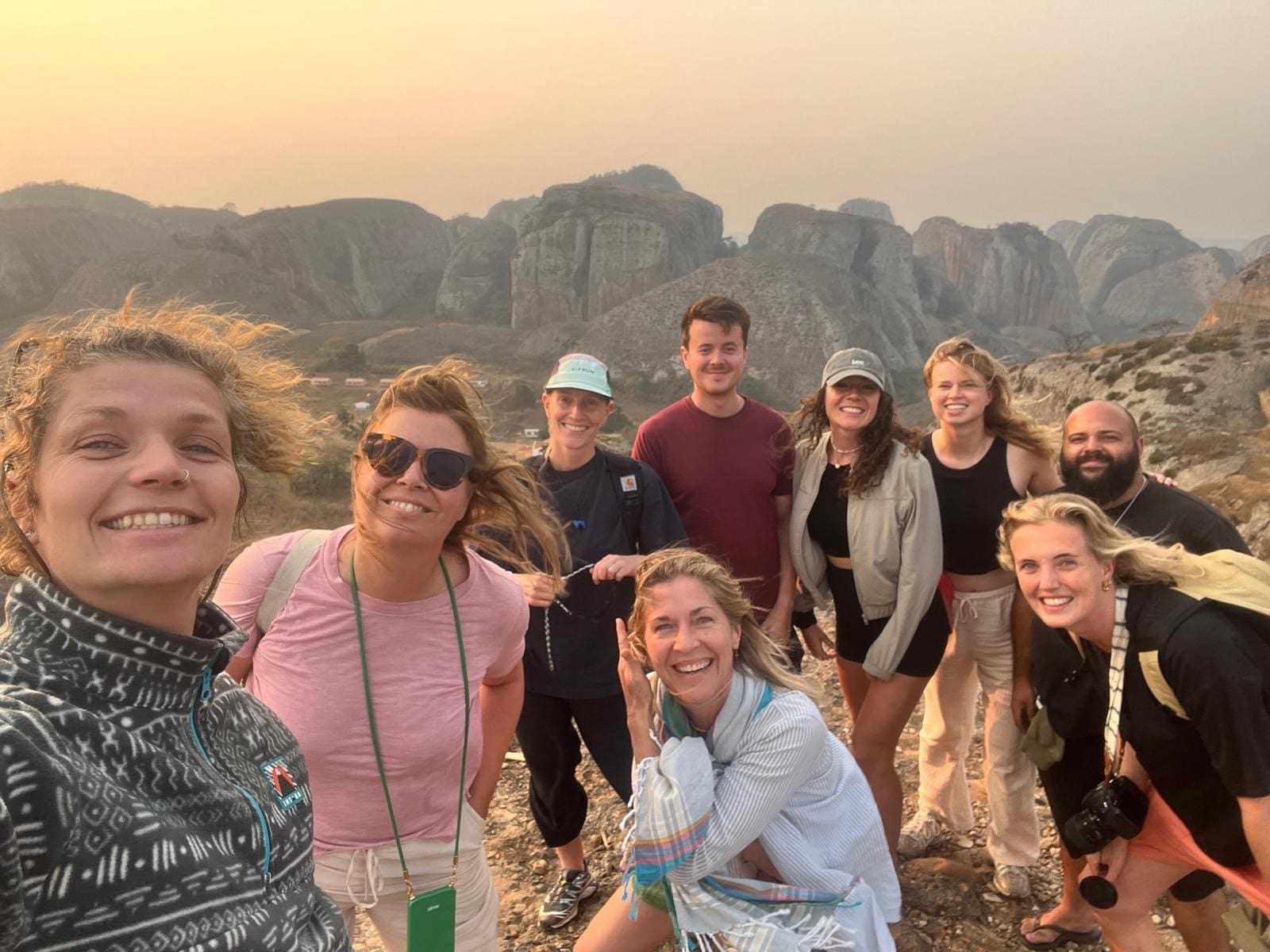 Een groep van tien lachende mensen poseert voor een foto buiten met rotsachtige bergen op de achtergrond tijdens zonsondergang. Sommigen dragen hoeden en vrijetijdskleding, en één persoon heeft een camera om zijn nek.
