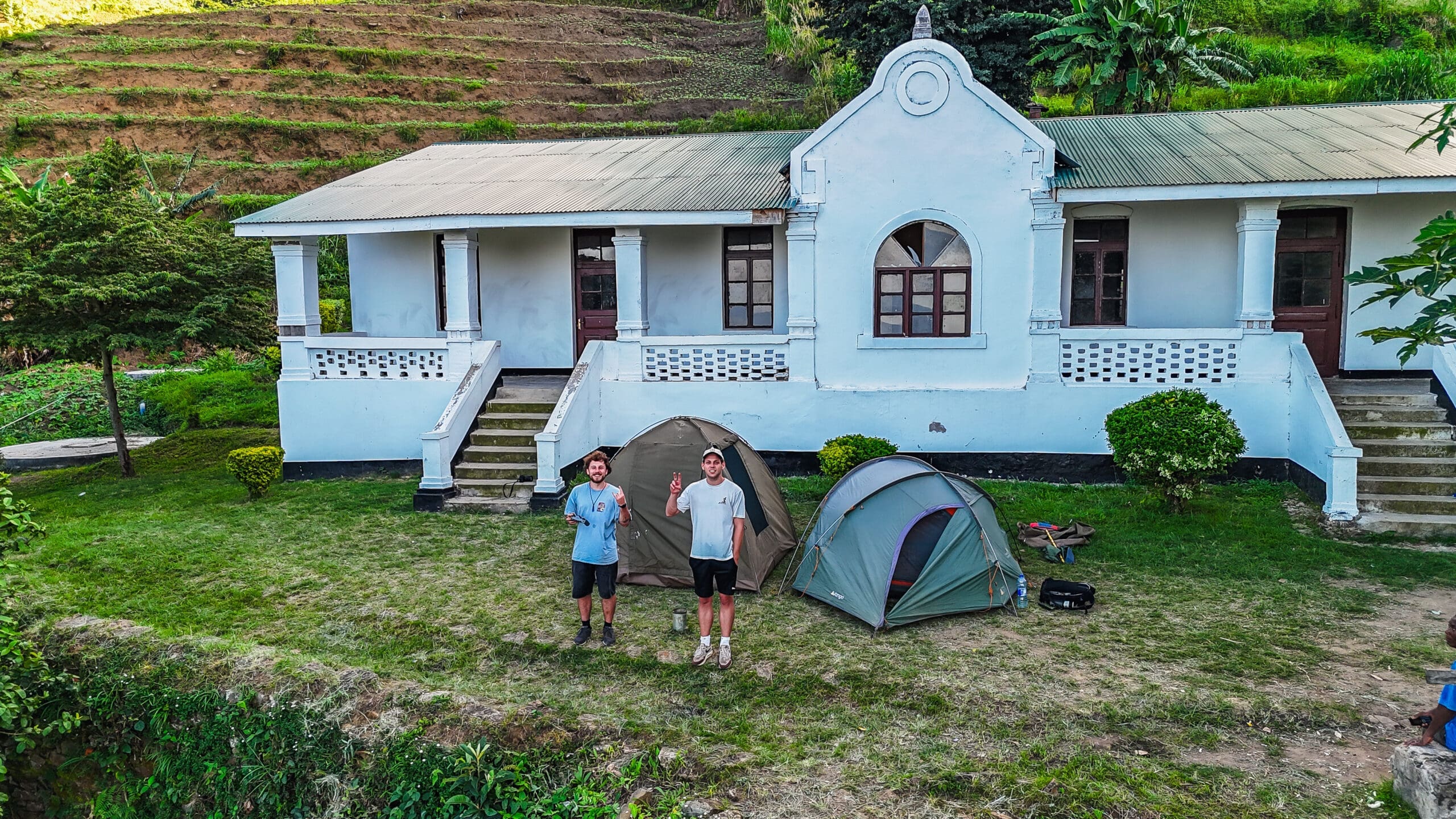 Twee mensen staan op gras naast twee tenten voor een wit gebouw met zuilen en boogramen, omringd door groen en terrasvormige heuvels op de achtergrond.