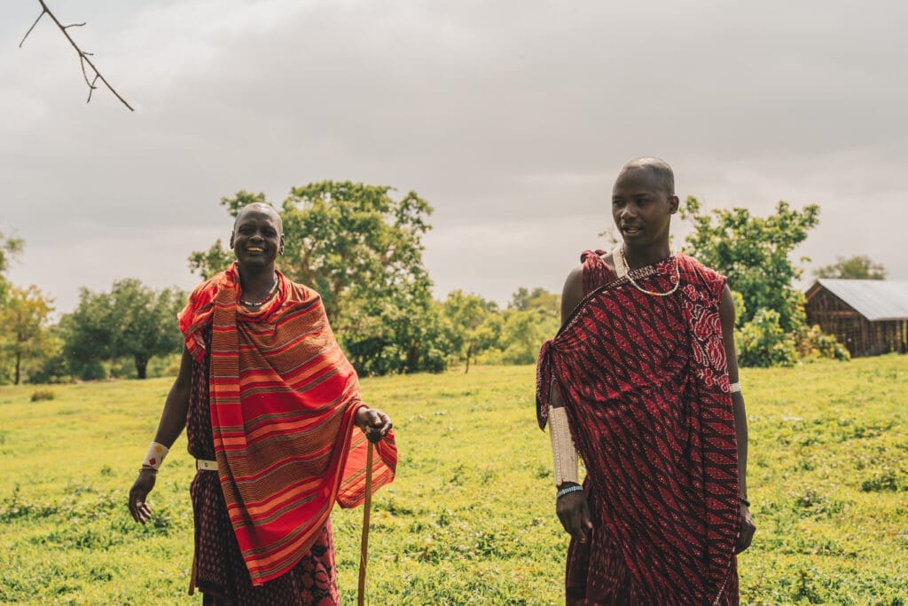 Twee mensen in traditionele Maasai kleding staan glimlachend buiten op groen gras, met bomen en een bewolkte lucht op de achtergrond. De een draagt een felrood gestreepte shuka en houdt een staf vast; de ander heeft een omslagdoek met patroon.